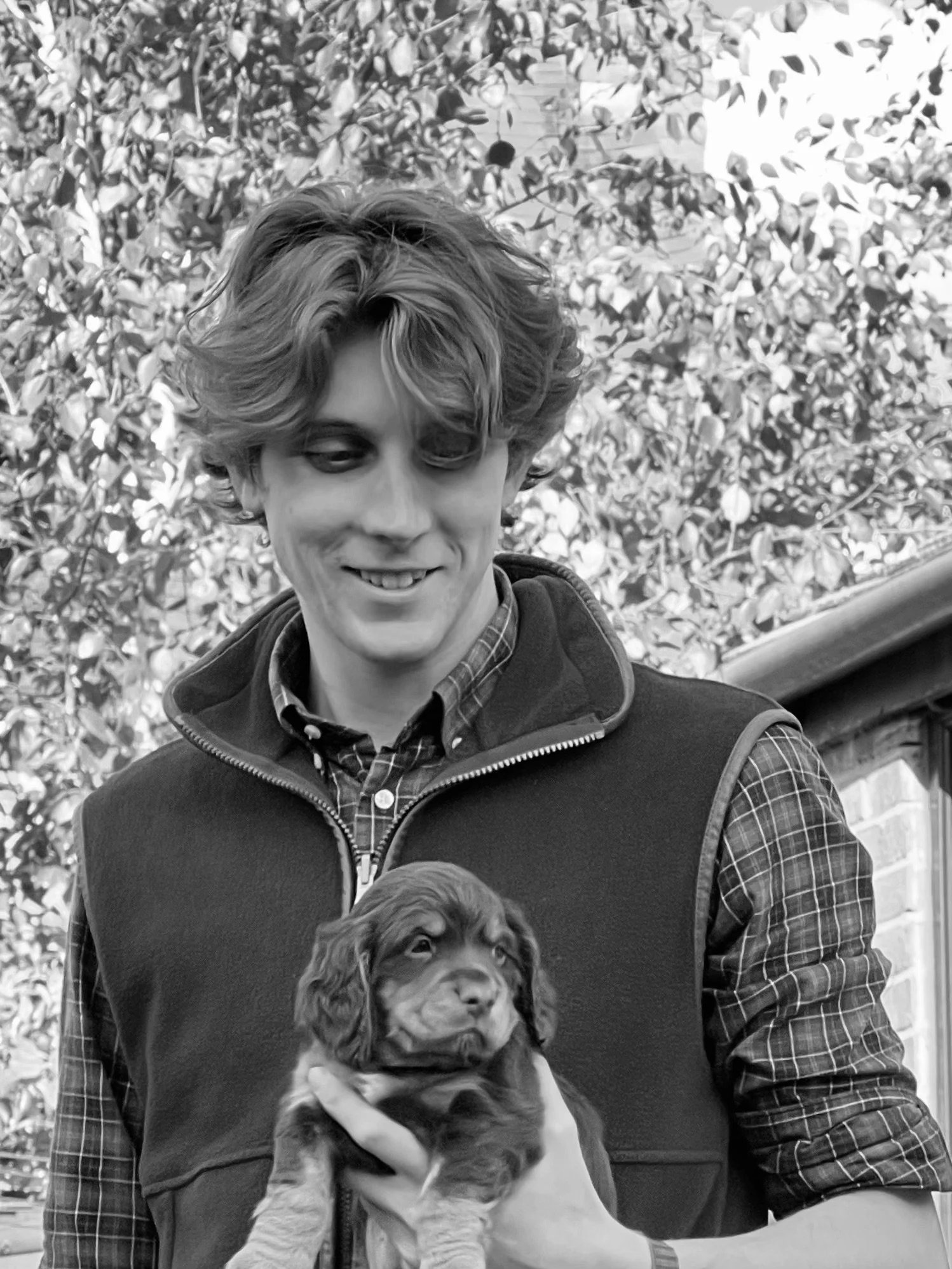A young man with wavy hair and dressed in a plaid shirt and vest holding a puppy outdoors with trees in the background.