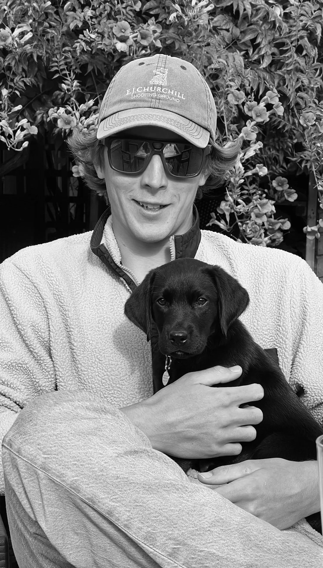 A man wearing sunglasses, a cap, and a fleece jacket sitting outdoors, holding a black puppy in his lap, with a background of leafy bushes.