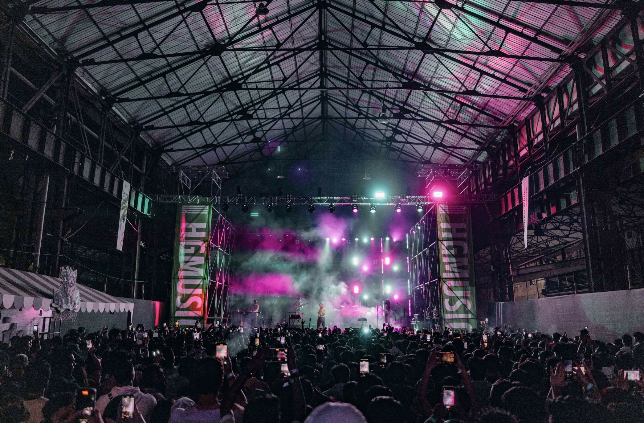 Concert or music event inside a large industrial-style venue with a high metal roof, colorful stage lighting, smoke effects, and a crowd of people taking photos and videos with their phones.