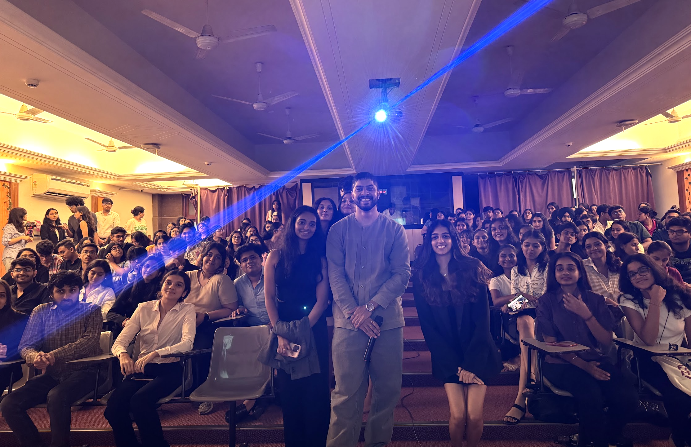 A large room filled with a diverse group of people, mostly young adults, attending a seminar or event. In the foreground, a man and three women are standing and smiling at the camera. The room has a ceiling with multiple fans, purple curtains, and a blue laser light projection.