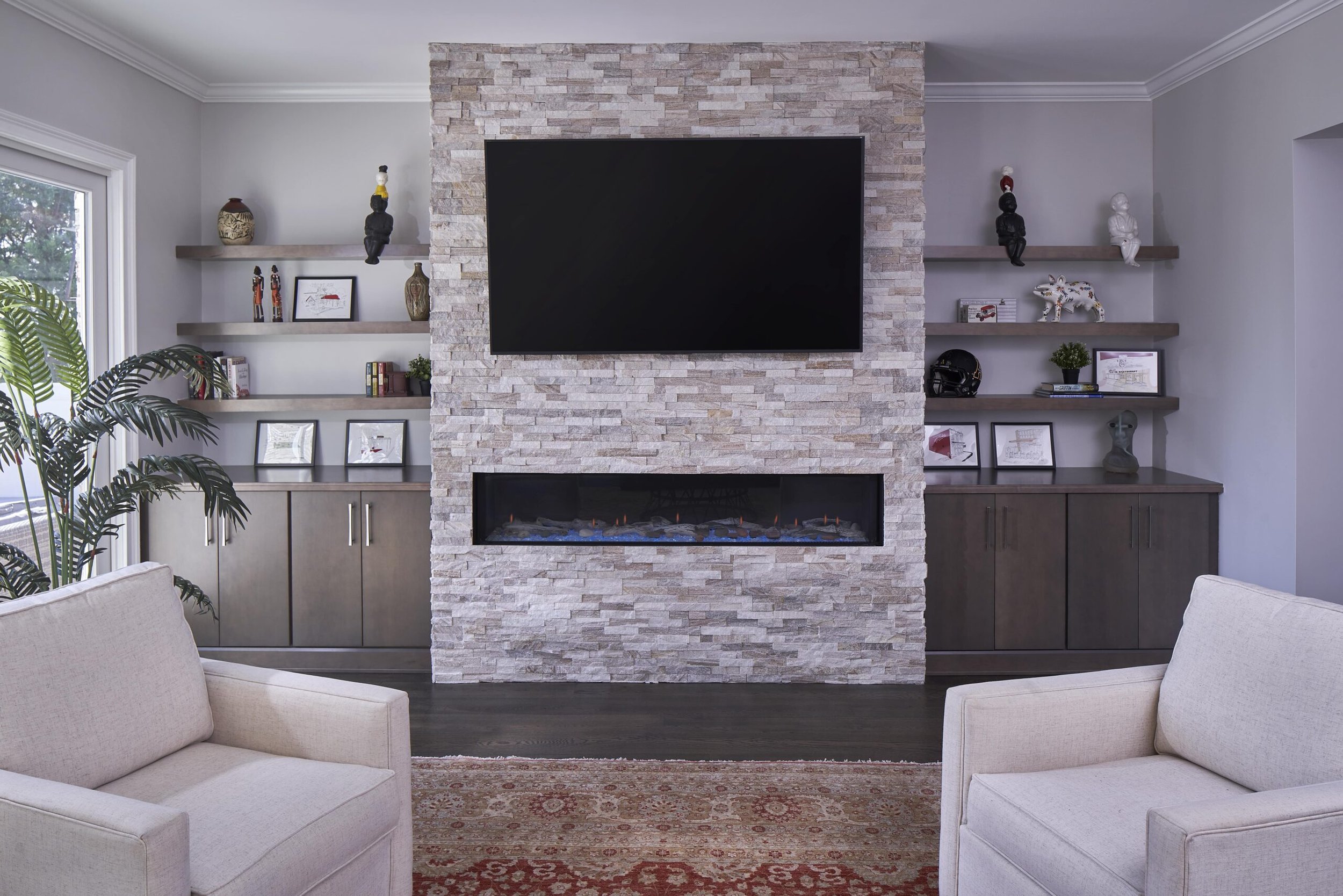 Living room with a stone fireplace, wall-mounted TV, built-in shelves with decorative items, beige armchairs, a patterned rug, a large plant near a window.