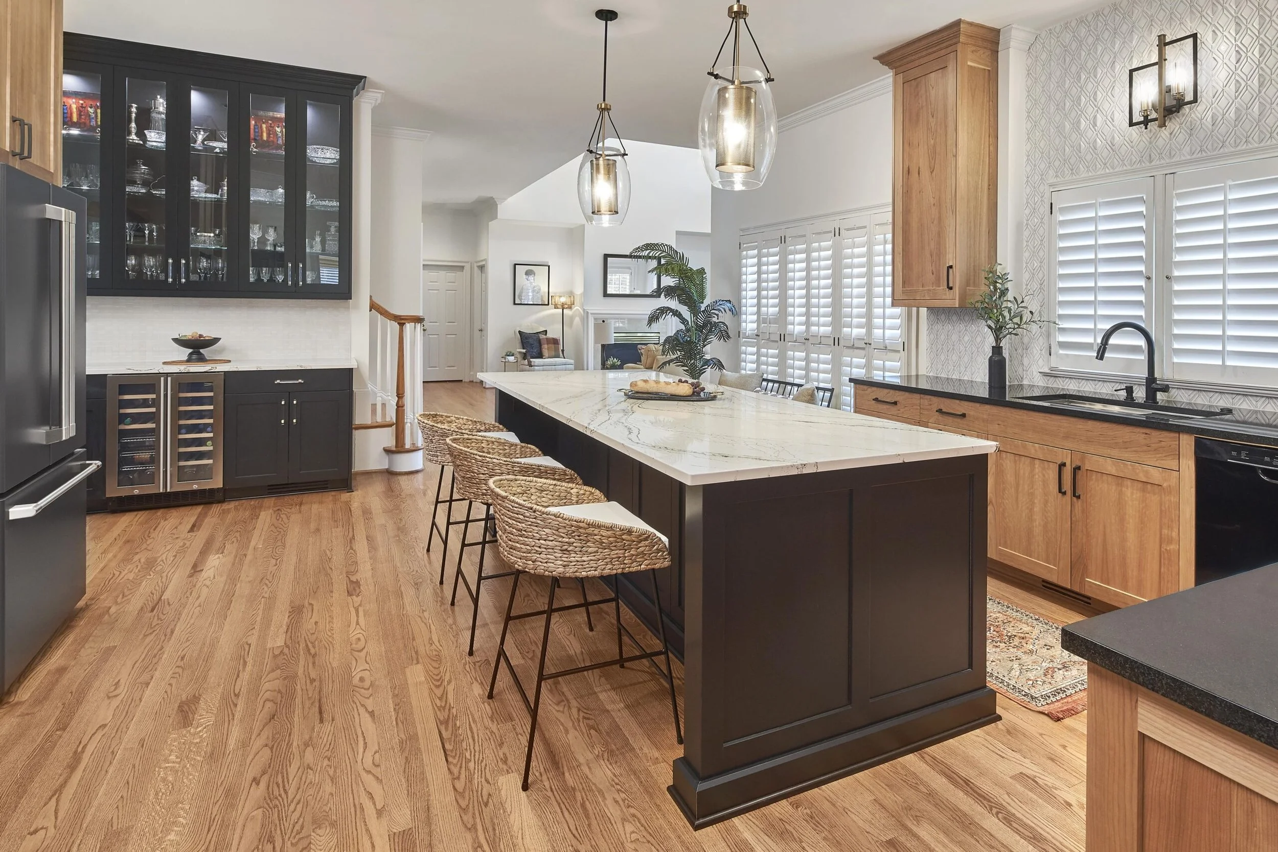 Modern kitchen with black and wooden cabinets, white marble island with four rattan bar stools, hardwood flooring, black appliances, presence of large windows with white shutters, and pendant lighting above the island.