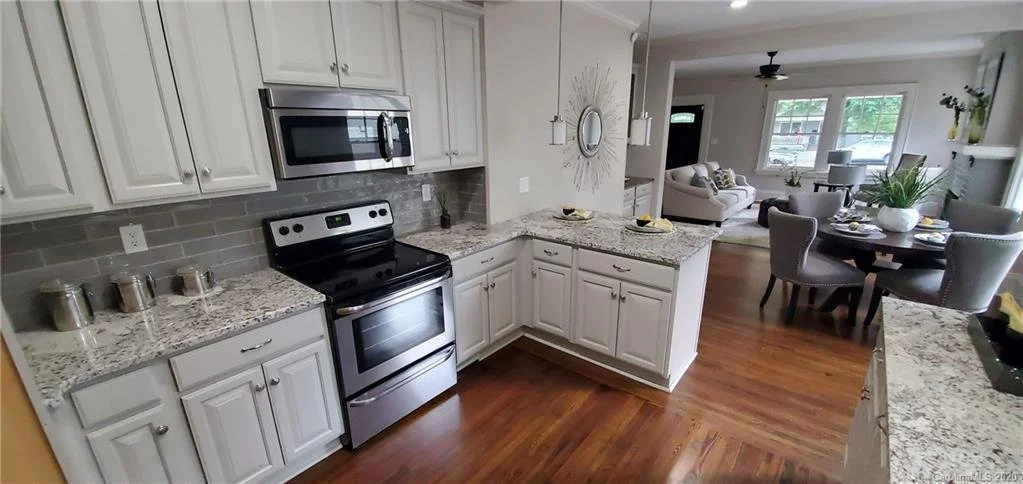Modern kitchen with white cabinets, stainless steel stove, granite countertops, and gray tile backsplash. Adjacent to the kitchen is a living area with a dining table, chairs, and a seating area with sofas and large windows.