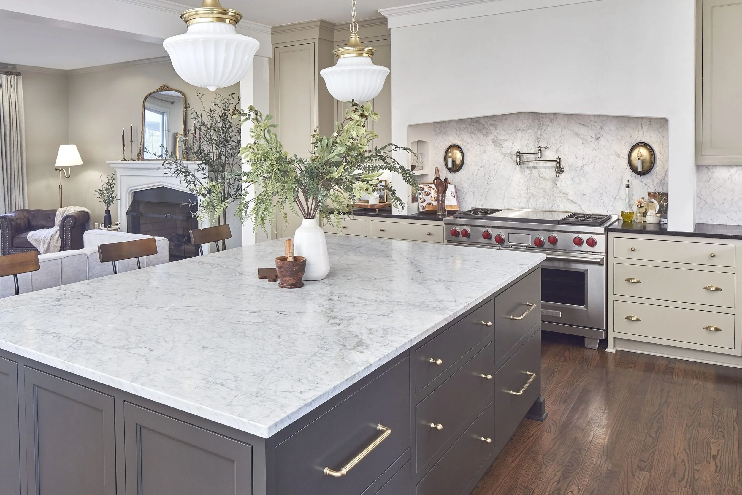 Modern kitchen with white marble countertops, beige cabinets, and stainless steel appliances. There are two white pendant lights above the island with a large white vase holding green foliage. The background shows a living room with a fireplace, a mi