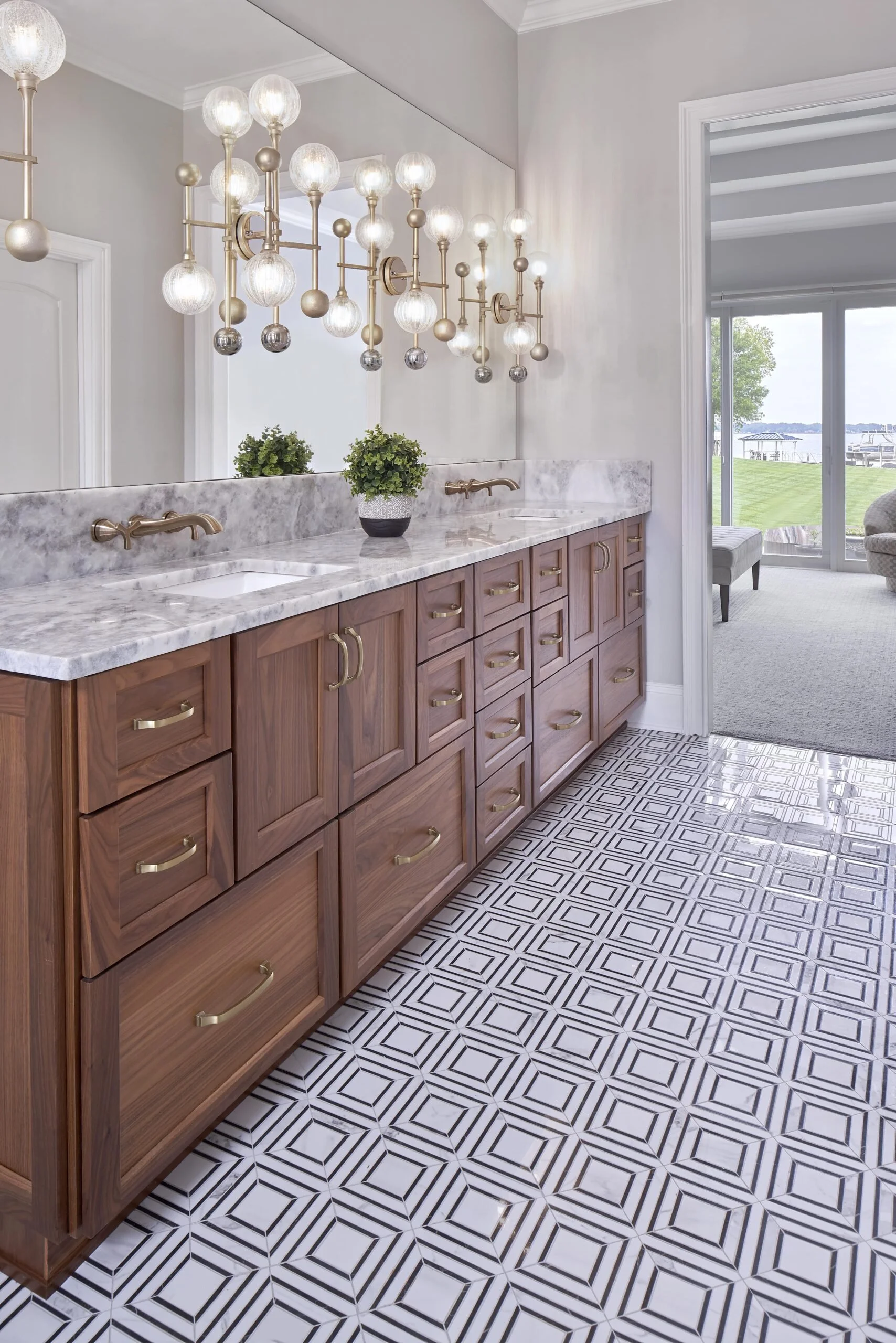 Bathroom with wooden cabinets, marble countertop, gold hardware, large mirror, decorative hanging light fixture, and a view of a backyard with lake in the background.