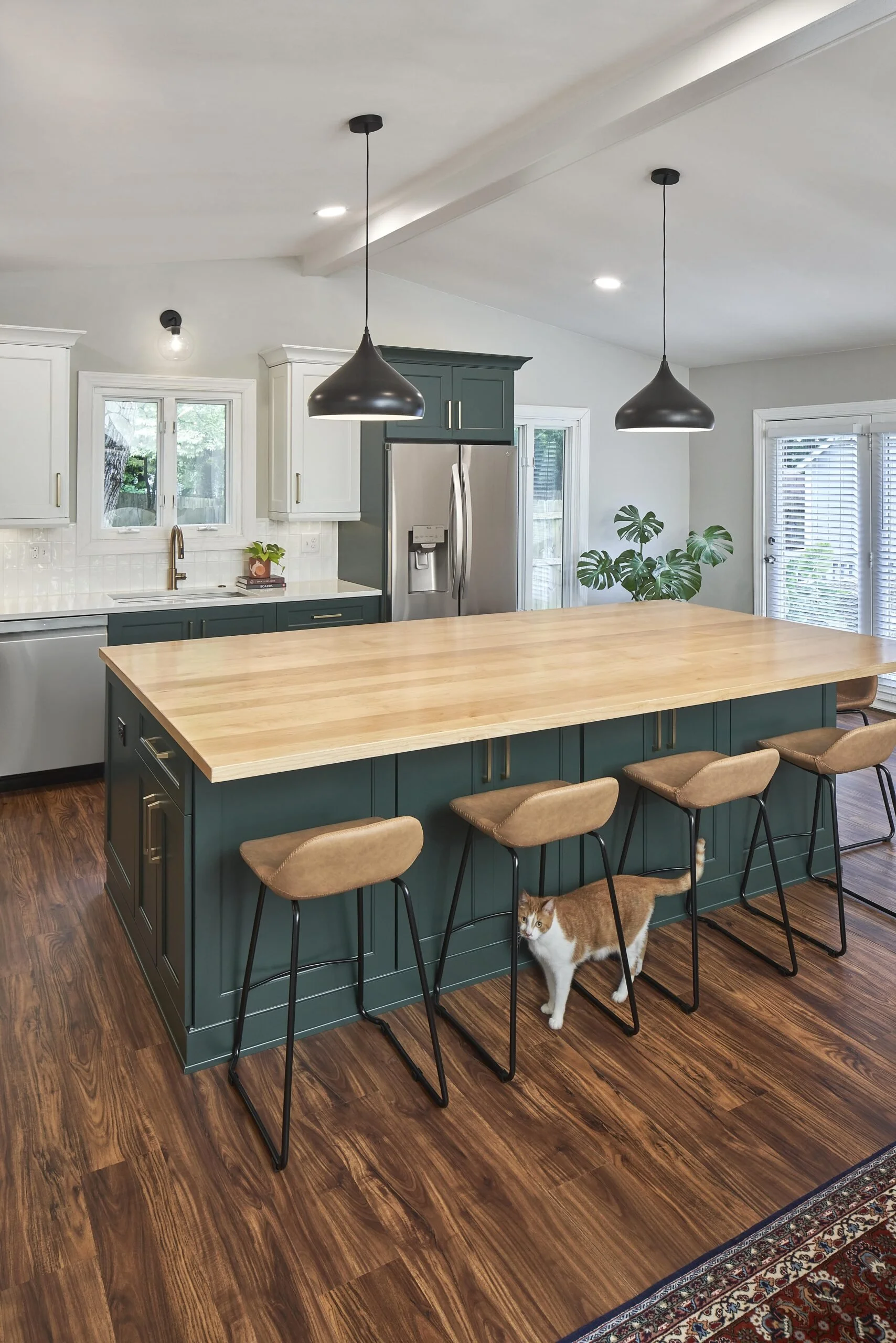 Modern kitchen with a large wooden island, white and teal cabinets, stainless steel appliances, hardwood floors, black pendant lights, a small cat under the island, and a plant near a sliding door.