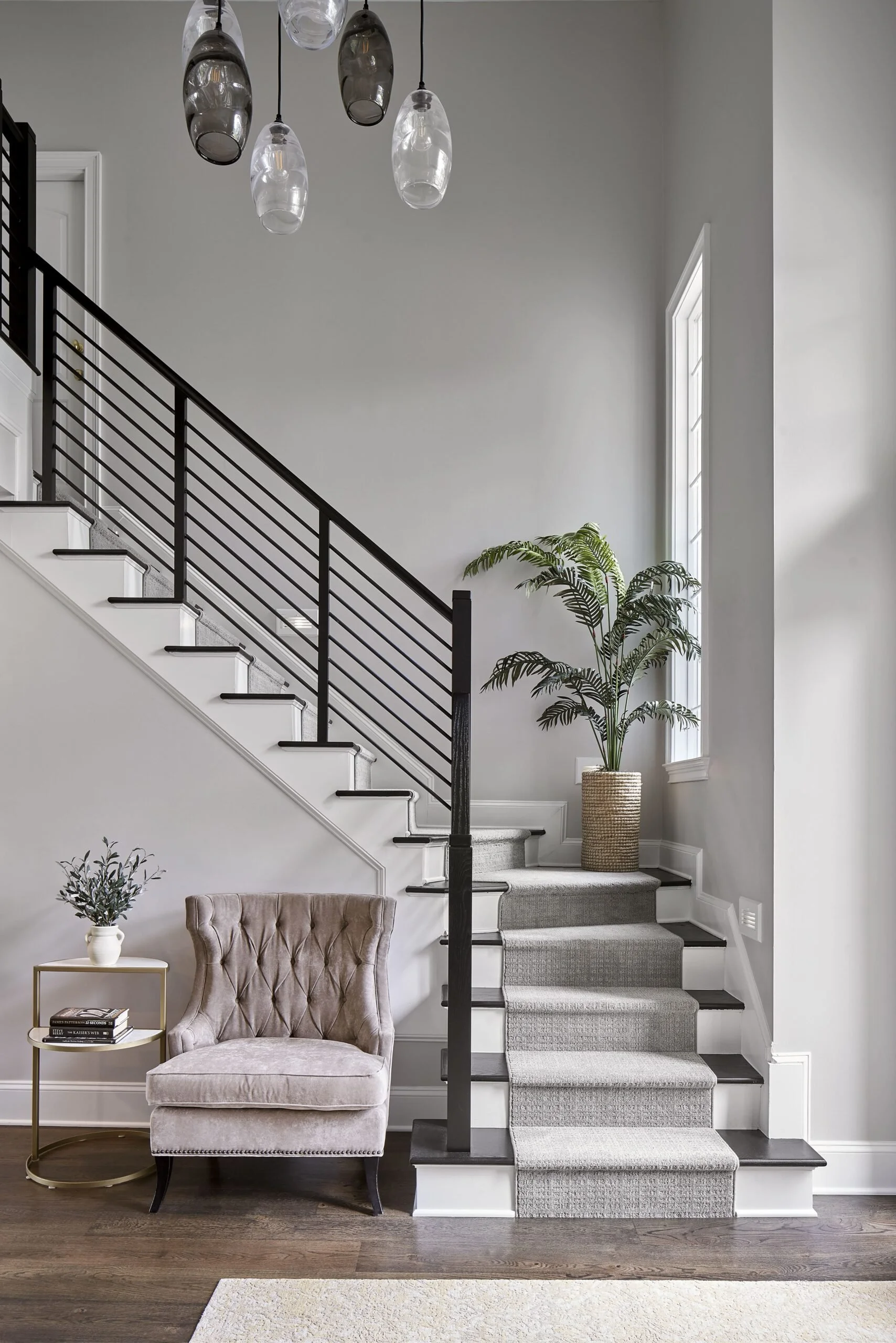 Interior of a modern living space featuring a staircase with black railing, a beige upholstered armchair, a side table with books and a plant, a large window with a potted plant by it, and a distinctive ceiling light fixture.