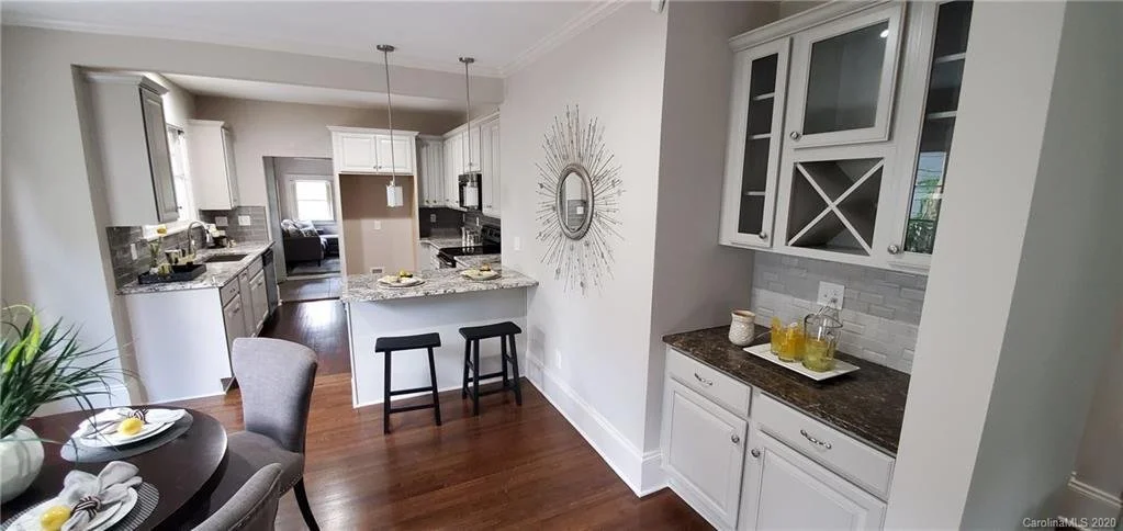 A view of an open kitchen and dining area with white cabinets, granite countertops, dark wood flooring, a small kitchen island with two black stools, and a round dining table with four gray upholstered chairs. There is a decorative round mirror on th