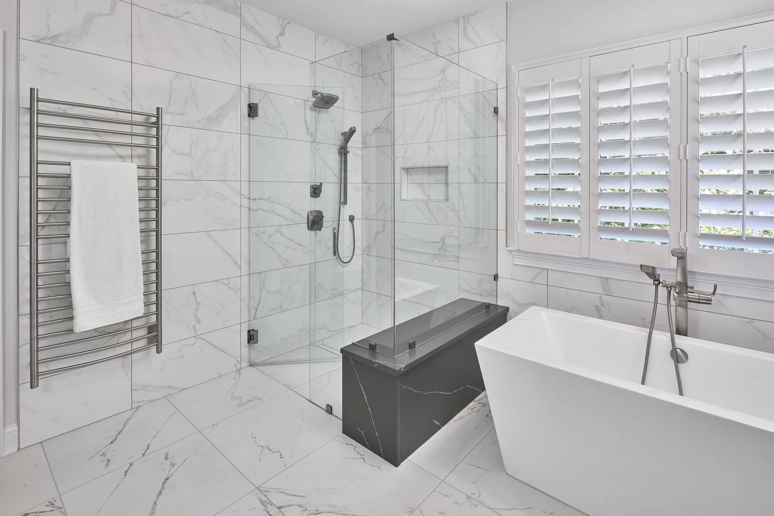 Modern bathroom with marble tiles, glass-enclosed shower, free-standing bathtub, window with shutters, and a towel radiator.