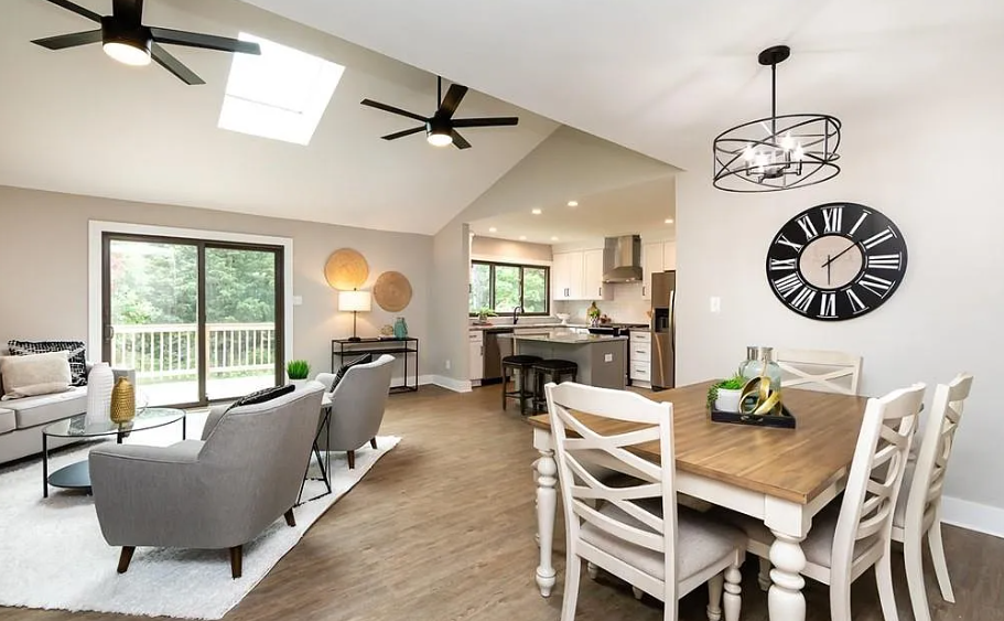 Open-concept living and dining area with gray couches, a white dining table, black and gray chairs, large wall clock, ceiling fans, and a view of the kitchen with white cabinets.