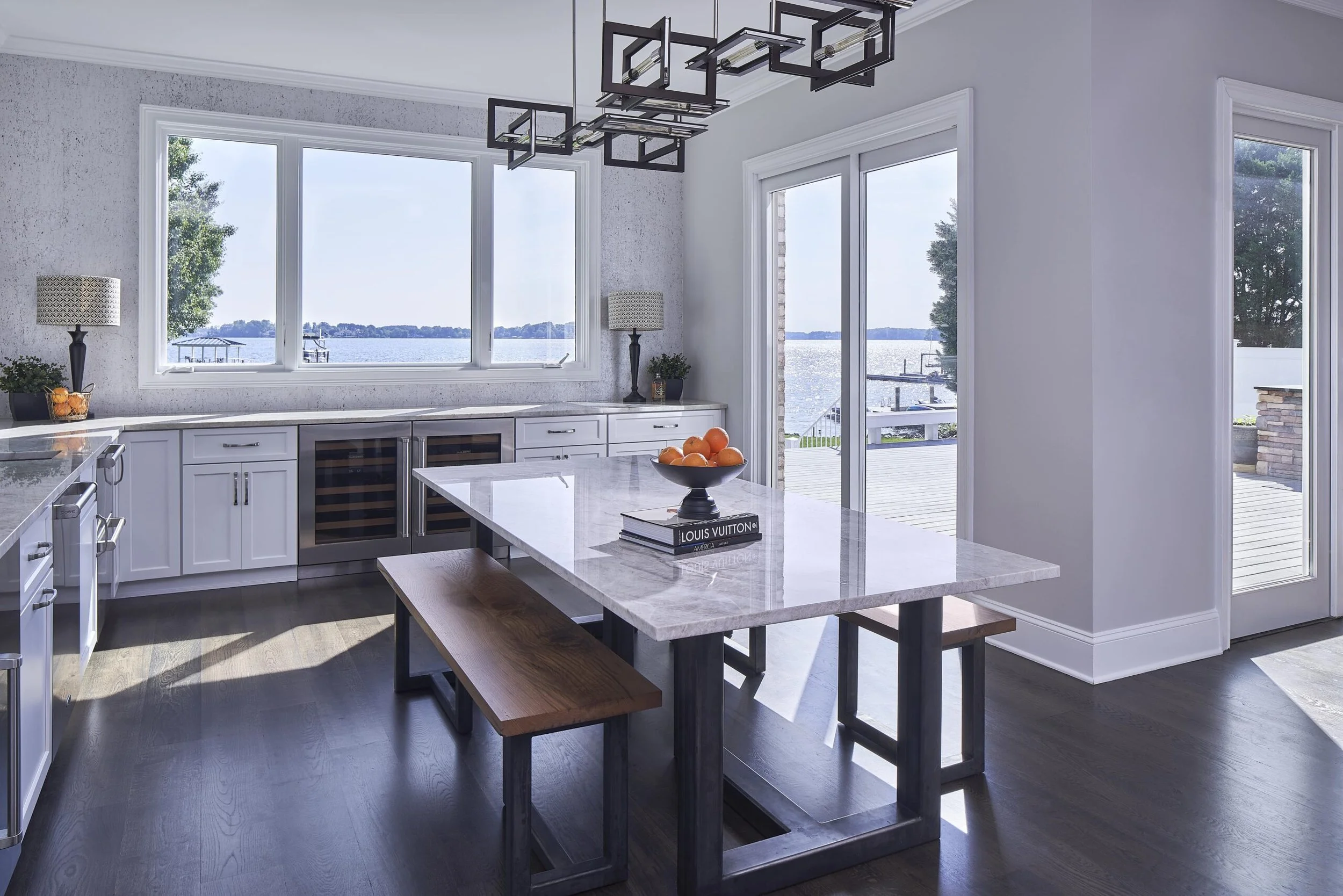 Modern kitchen with large windows overlooking a lake, a marble island with a bowl of oranges, and outdoor patio access.