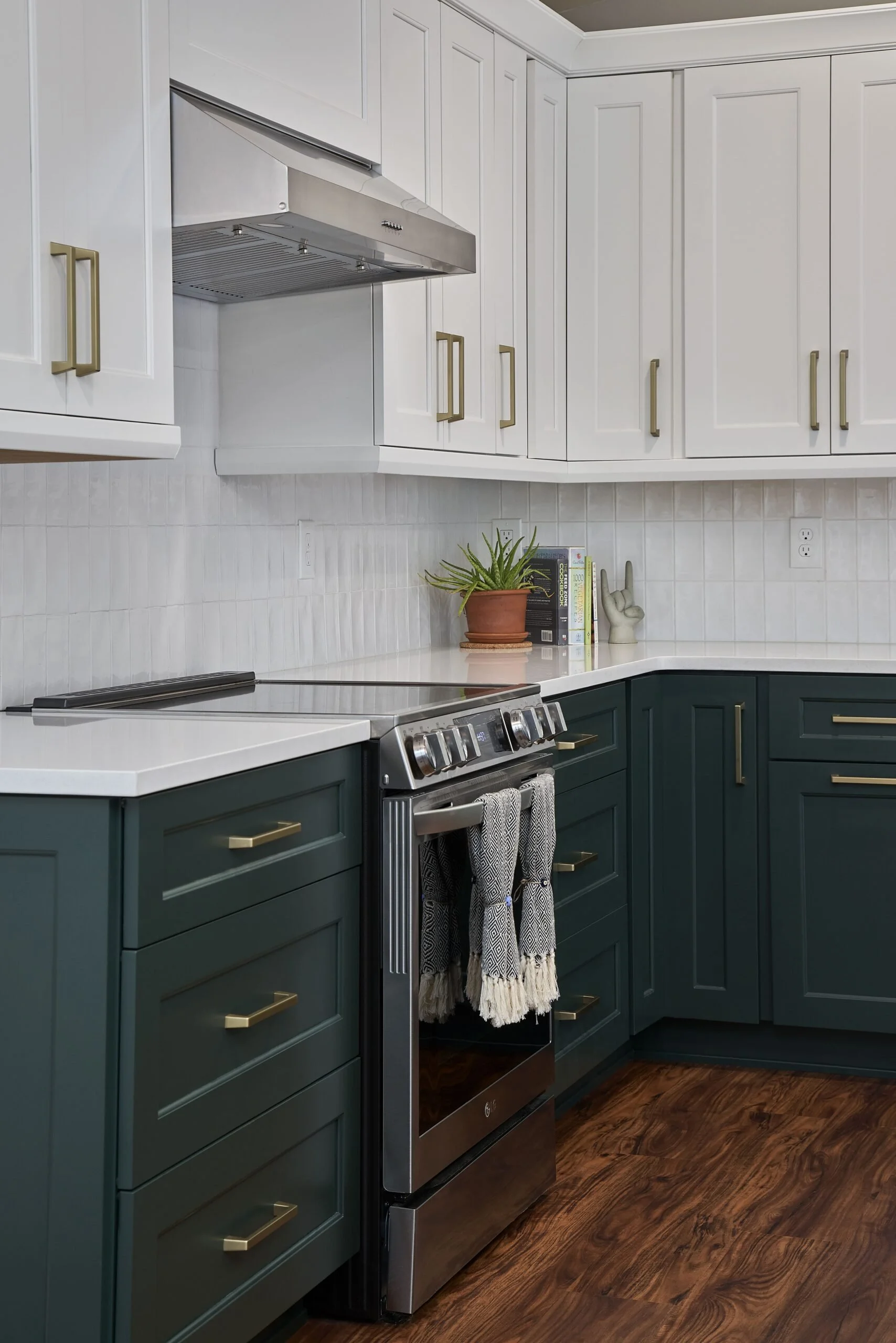 A kitchen with white upper cabinets and dark green lower cabinets, stainless steel stove, white countertops, a potted plant and books on the counter, hardwood flooring, and a range hood.