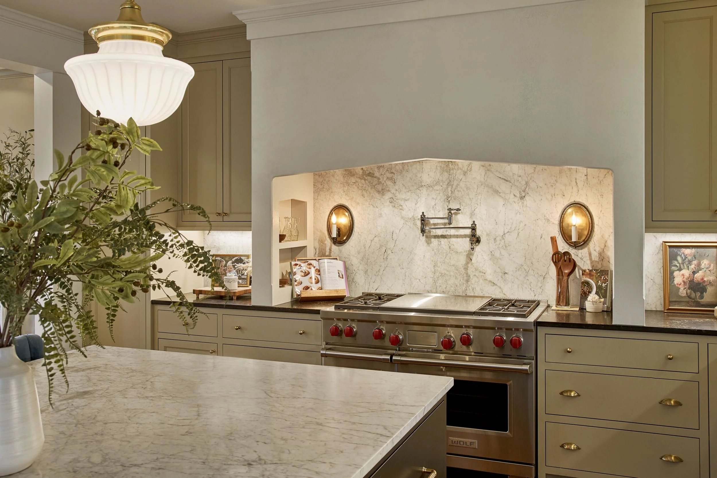 Kitchen with marble backsplash, modern stove with red knobs, green cabinets, and a marble island with a vase of greenery.