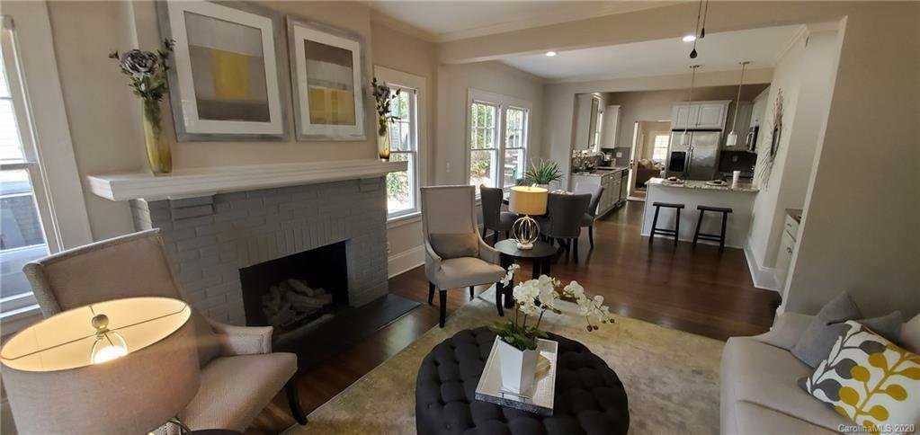 Living room with a fireplace, armchairs, a sofa with yellow and gray pillows, a round ottoman with flowers, a yellow lamp, a dining table with chairs, and a view into the kitchen.