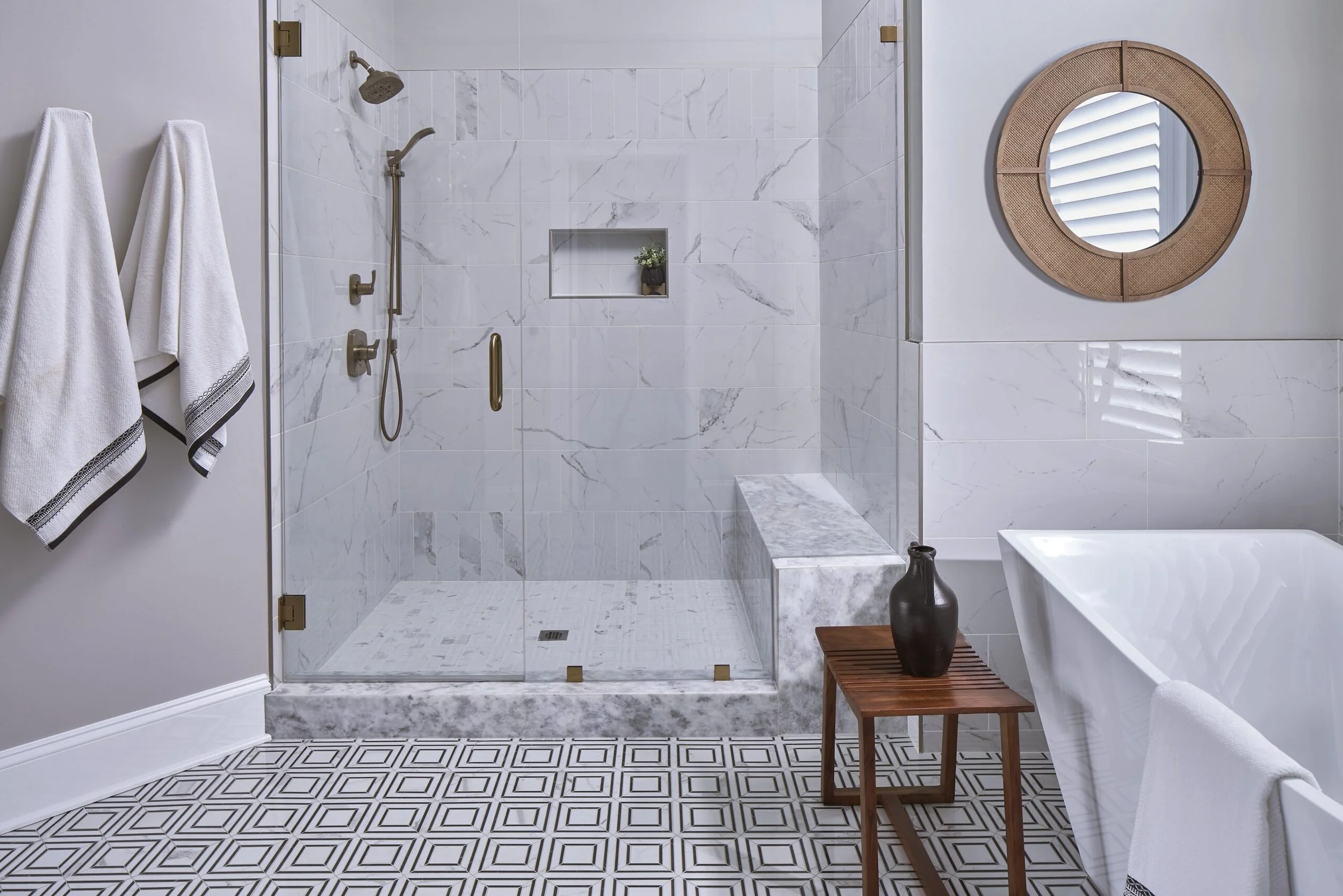 Modern bathroom with a glass walk-in shower, white marble tiles, a round wicker-framed mirror, a white bathtub, and patterned floor tiles. Towel hooks with white towels and a small wooden table with a black vase are also visible.