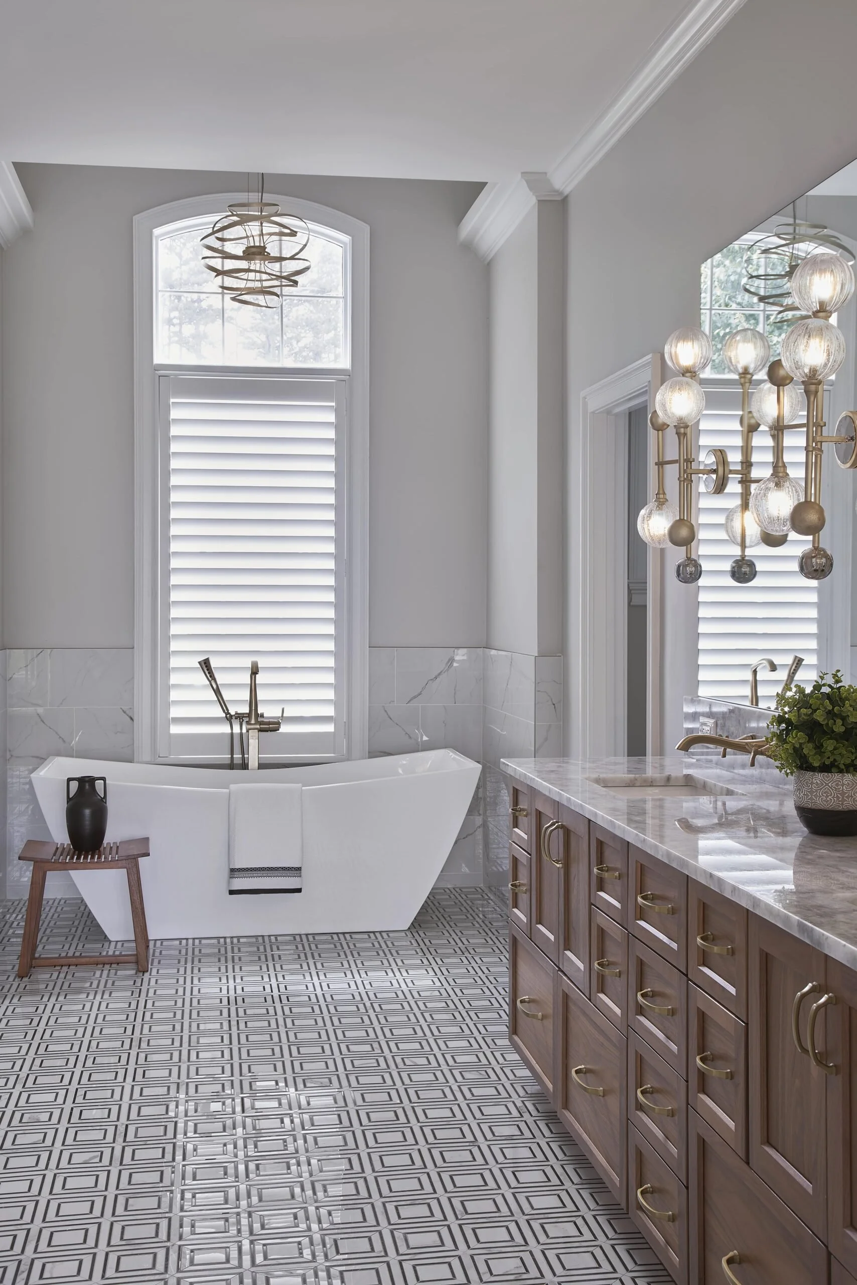 A modern bathroom featuring a white bathtub with a floor-mounted faucet, a wooden side table with a black vase, patterned tile flooring, a marble countertop vanity with brass hardware, a large mirror, decorative chandeliers, and plantation shutters o