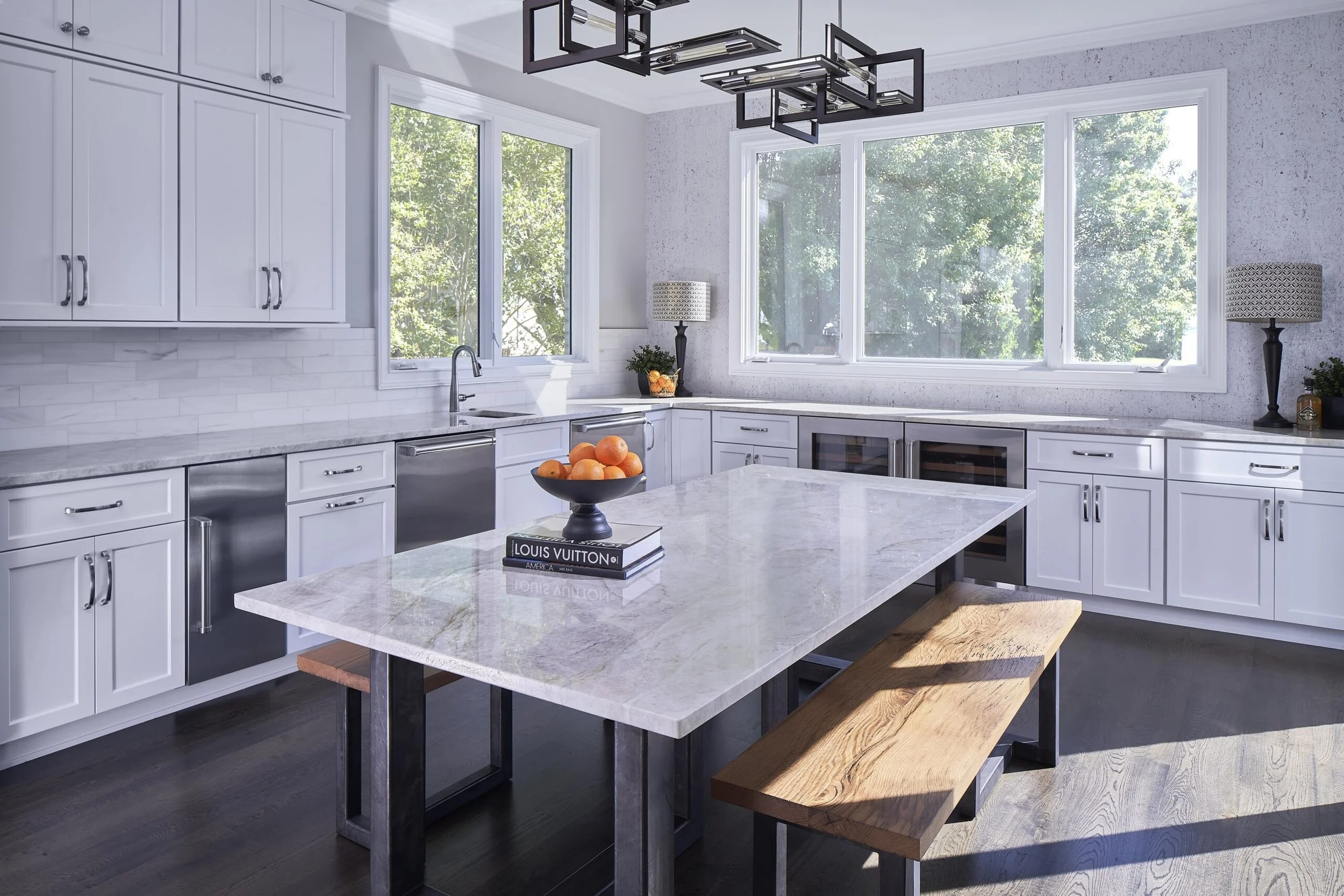Modern kitchen with large windows, white cabinets, marble countertops, and a kitchen island with a wooden bench. There are two table lamps, a bowl of oranges, and a few books on the island.