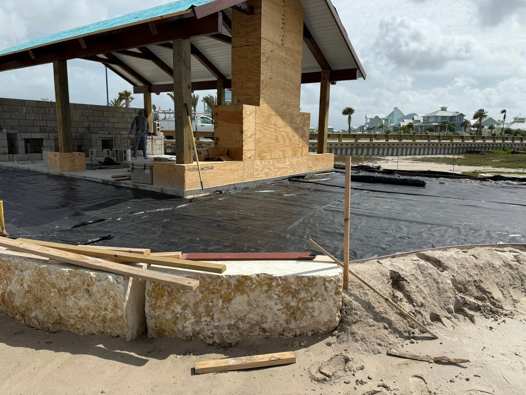 Construction site of a beach house with a wooden structure and partial wall, black waterproofing on the ground, and construction materials such as wood and concrete blocks, under a cloudy sky.