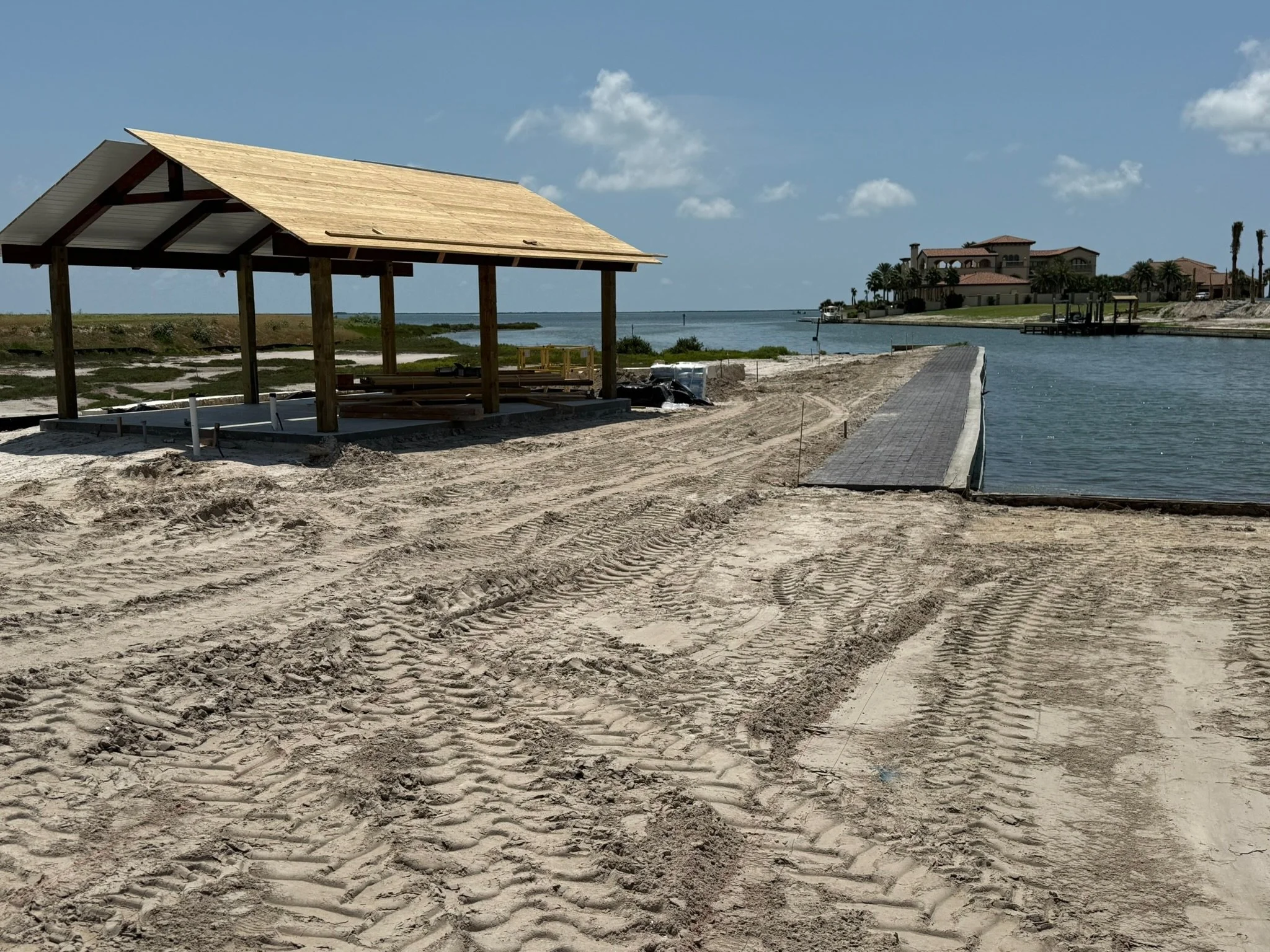 Construction site on a sandy beach with a wooden pavilion and a paved pathway leading to a body of water, with residential houses in the background under a partly cloudy sky.