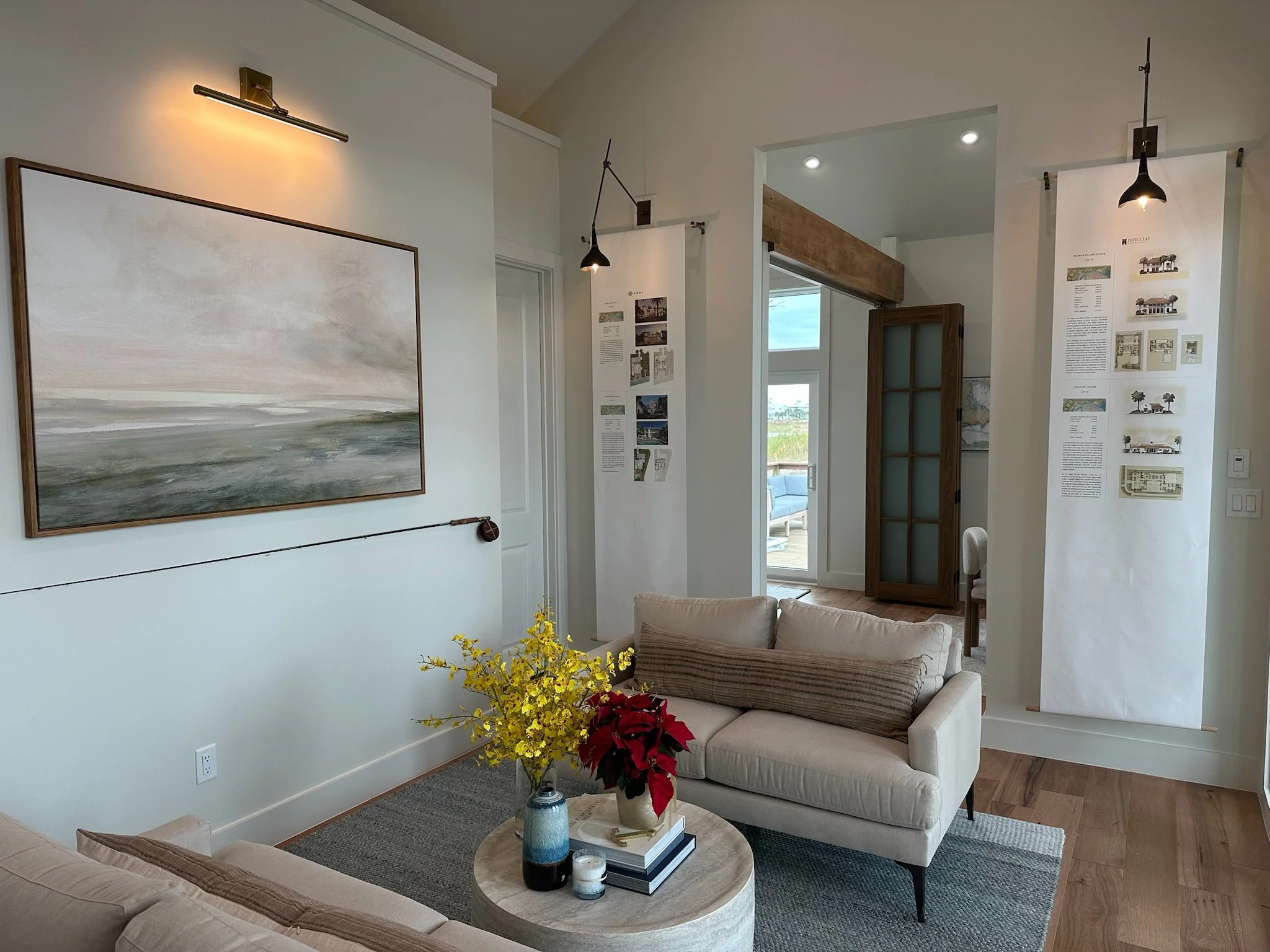 Living room with beige sofas, a round coffee table with flowers, a large landscape painting on the wall, and informational displays on the walls. A doorway leads to an outdoor patio visible through a glass door.