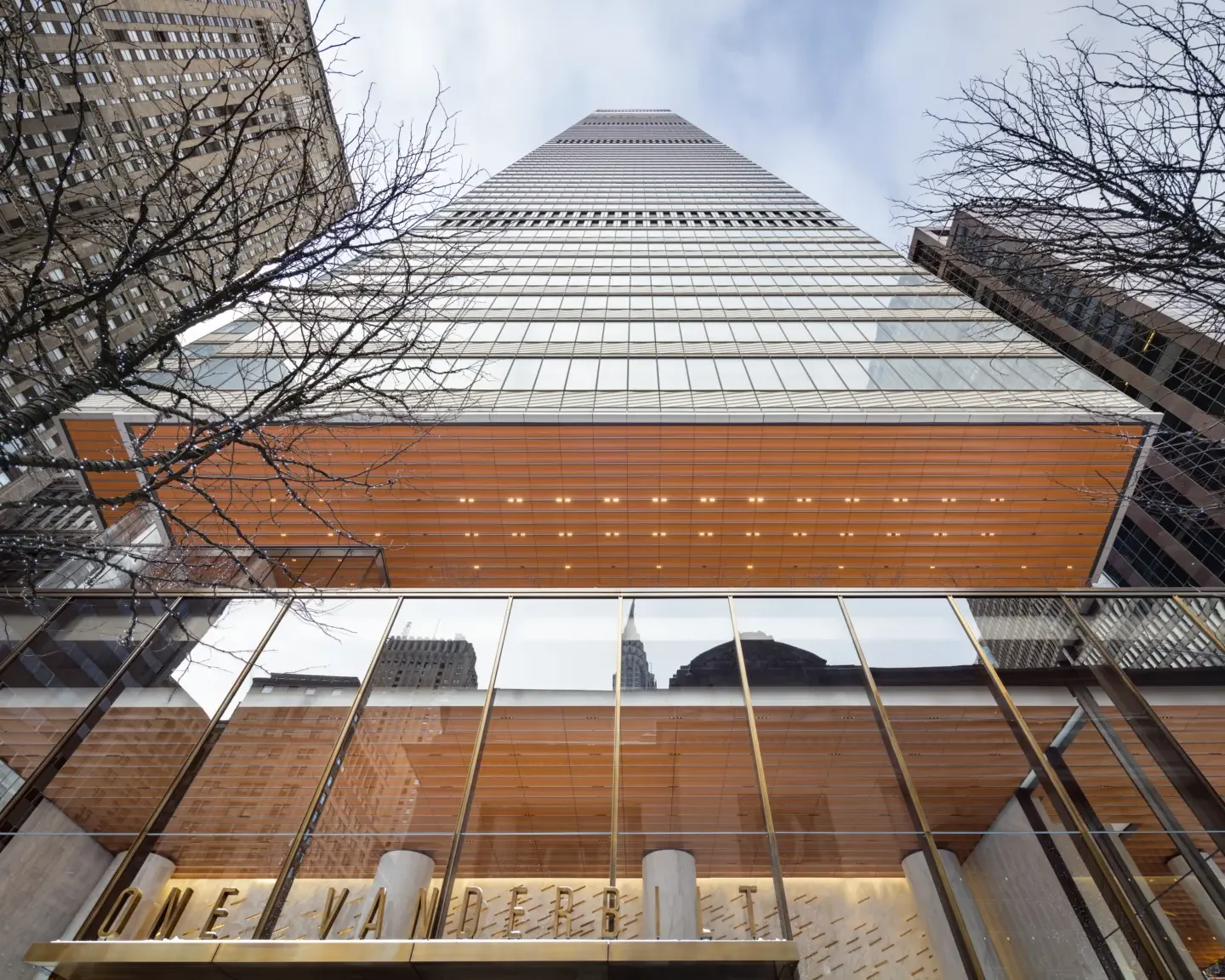 Looking up at a tall skyscraper with a glass facade, orange accents, and surrounding buildings, with leafless trees in the foreground.