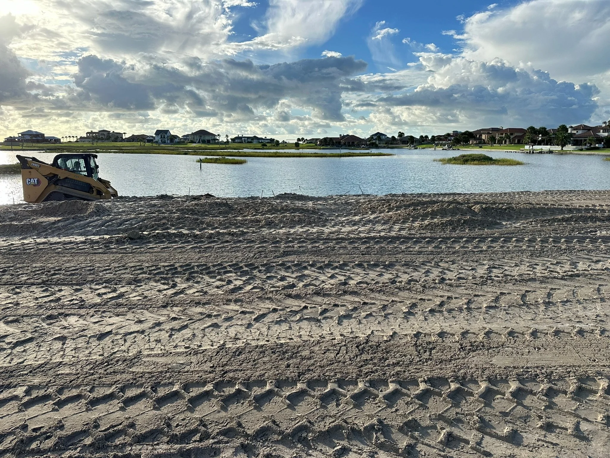 Construction equipment, a small bulldozer, on sandy ground near a water body with houses and cloudy sky in the background.