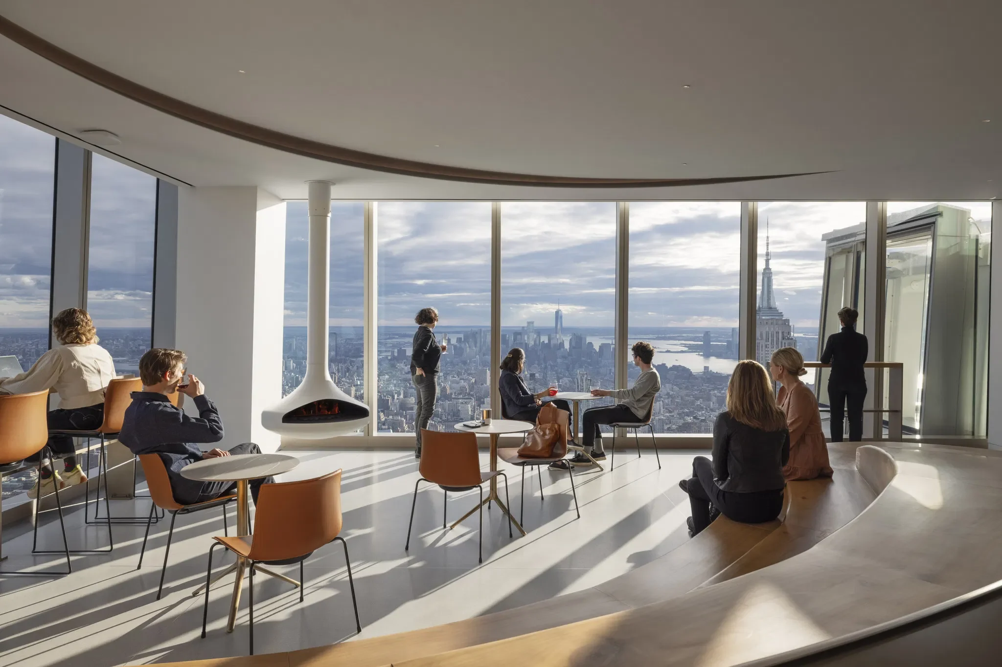 People sitting and standing in a modern lounge with large windows overlooking a city skyline, including the Empire State Building.