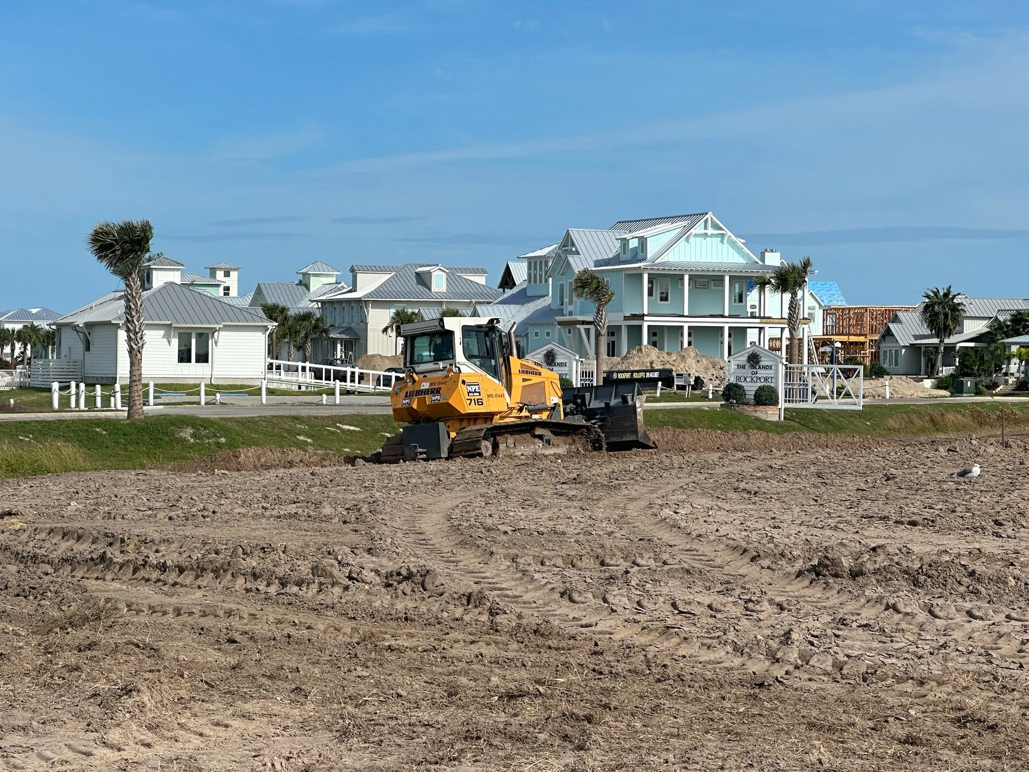 Construction excavator working on a beach with colorful houses in the background.