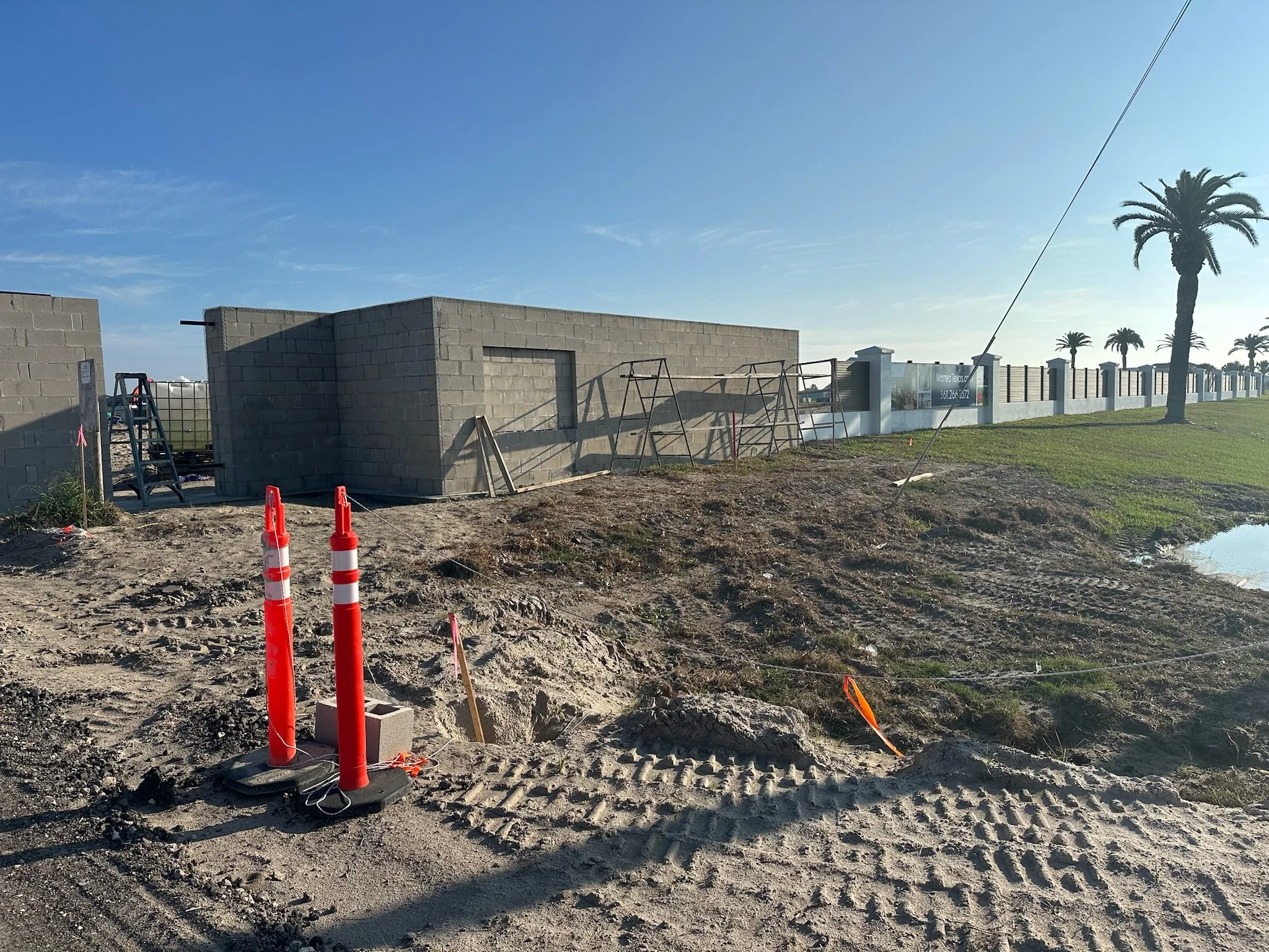 Construction site with a small concrete building, orange safety cones, dirt ground with tire tracks, green grass, trees, and a white fence under a clear blue sky.