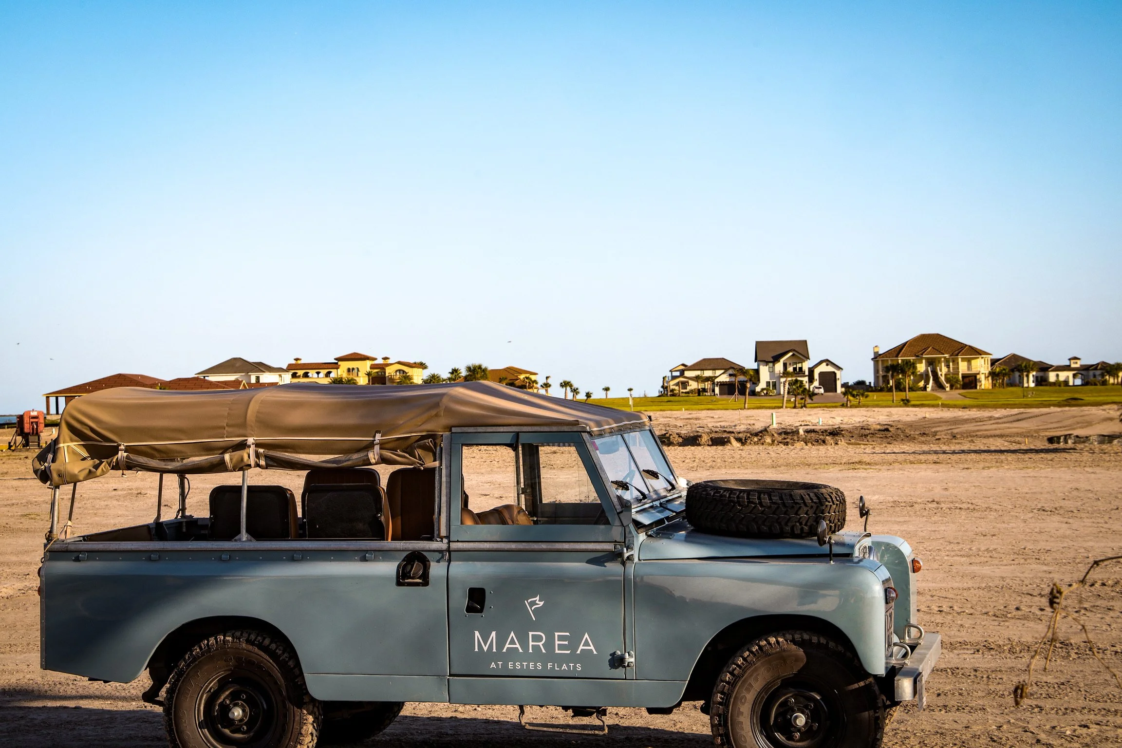 Vintage off-road vehicle with a spare tire on the hood, parked on a sandy beach, with residential houses in the background under a clear blue sky.