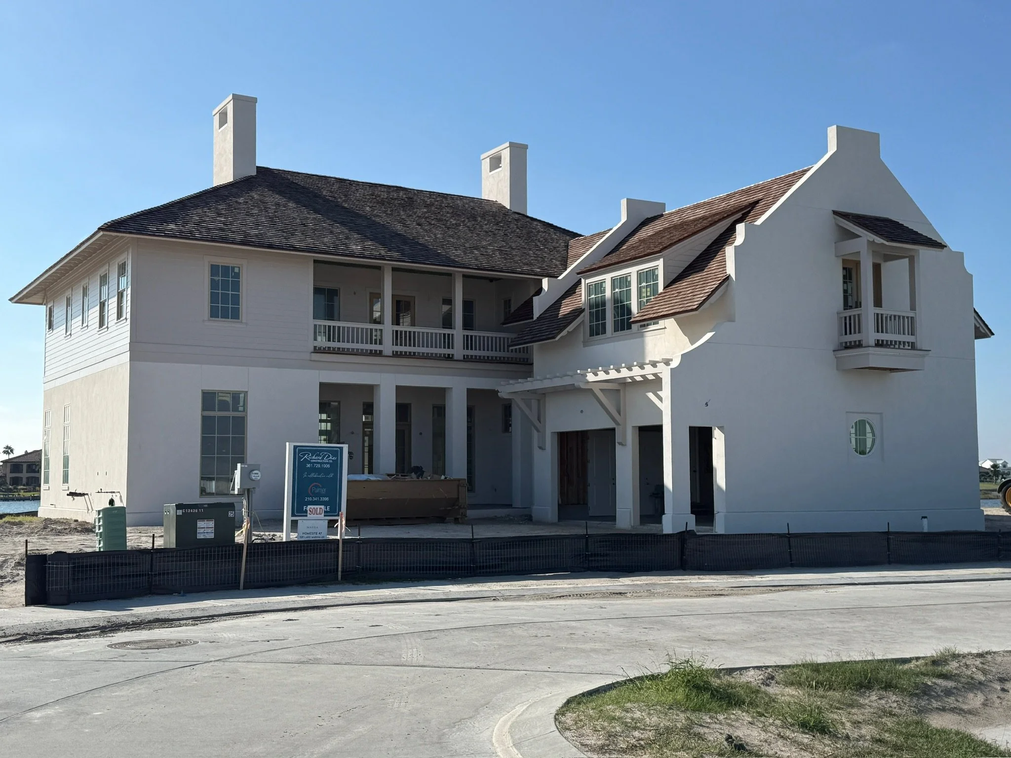 A large, white, multi-story house under construction with scaffolding and construction signs in front, facing a paved road, with a clear blue sky in the background.