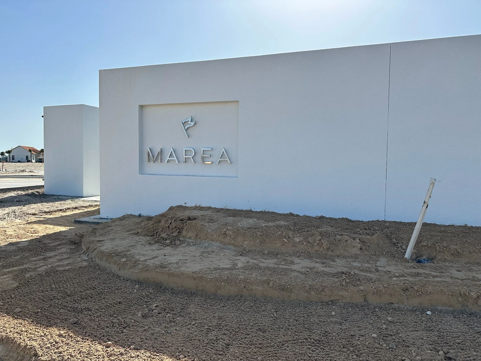 White exterior wall with a sign that reads 'MAREA' and a decorative emblem above it, set in a construction site with dirt and a partially installed light pole in the foreground.