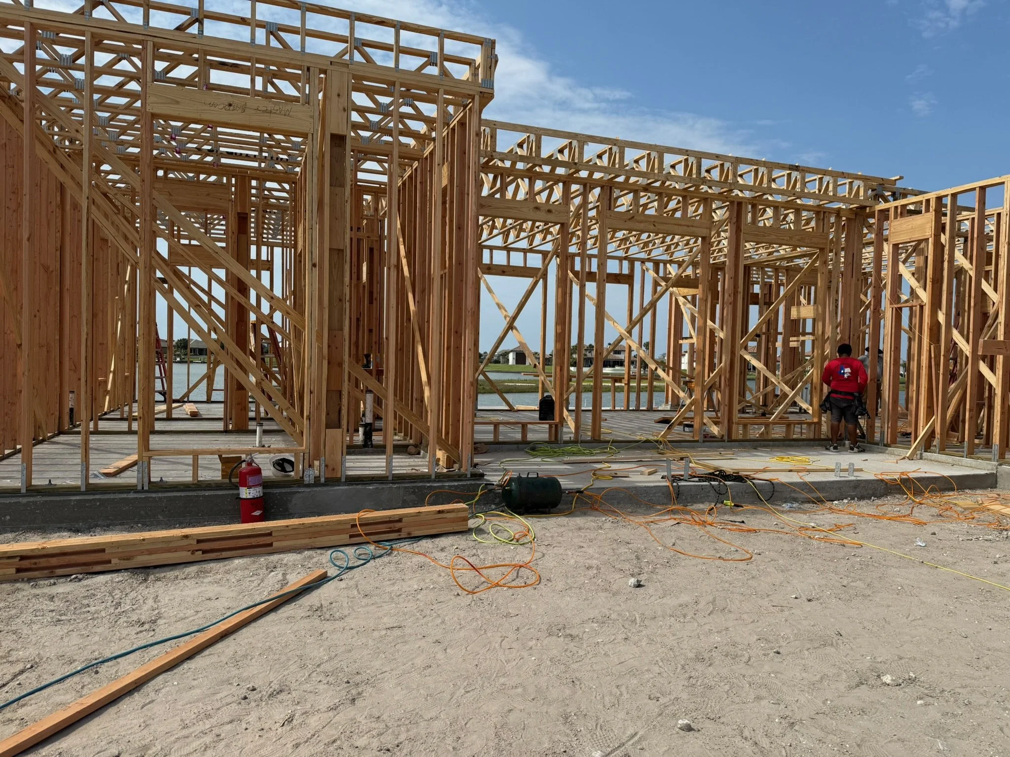 Wooden framework of a house under construction with construction workers on site. Building materials and tools are scattered on the sandy ground, and the sky is partly cloudy.