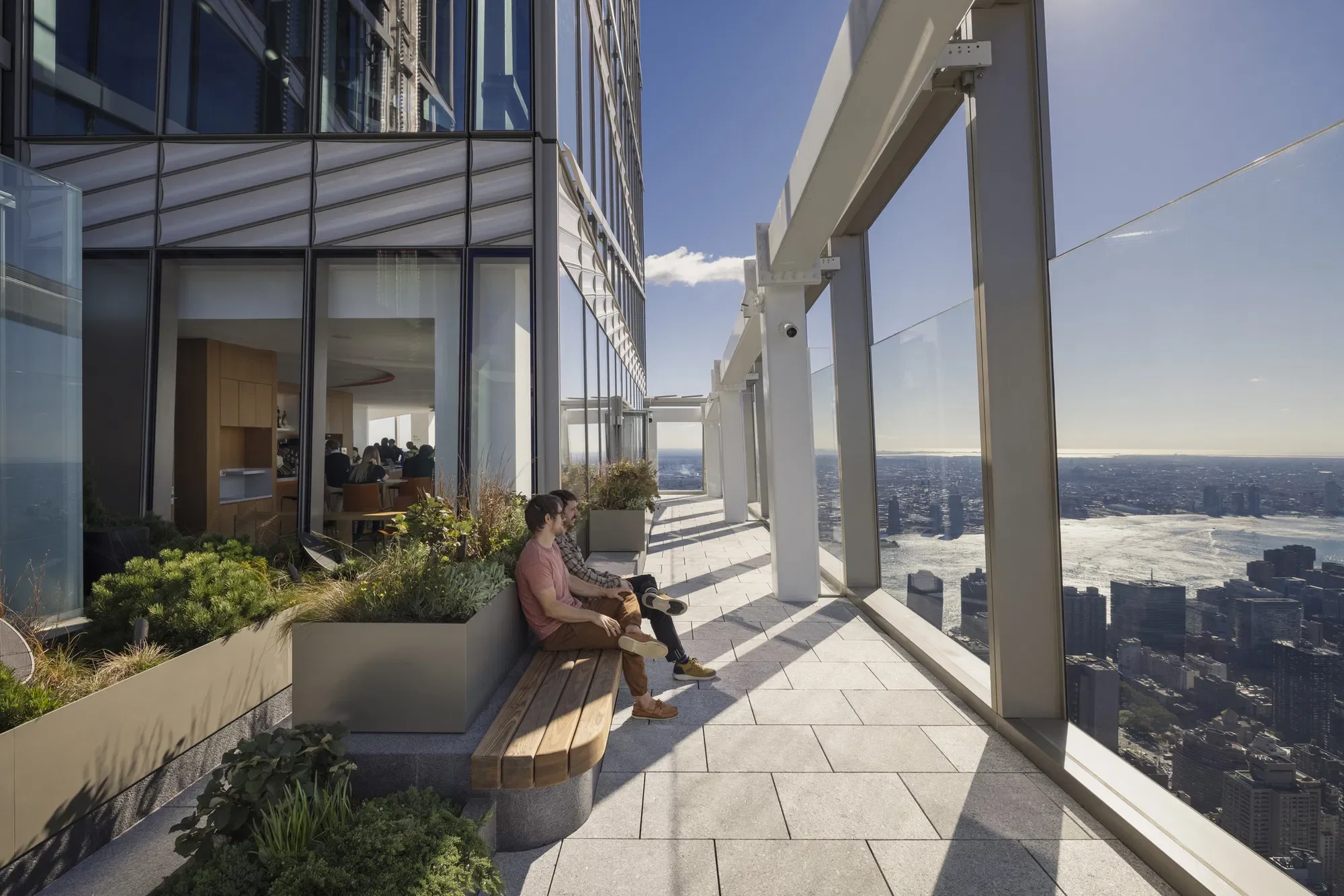 People sitting on a balcony with glass walls overlooking a city skyline and river on a sunny day.