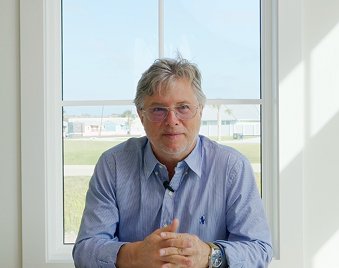 A man with gray hair and glasses sitting at a table in front of a large window, with a bright outdoor scene visible behind him.