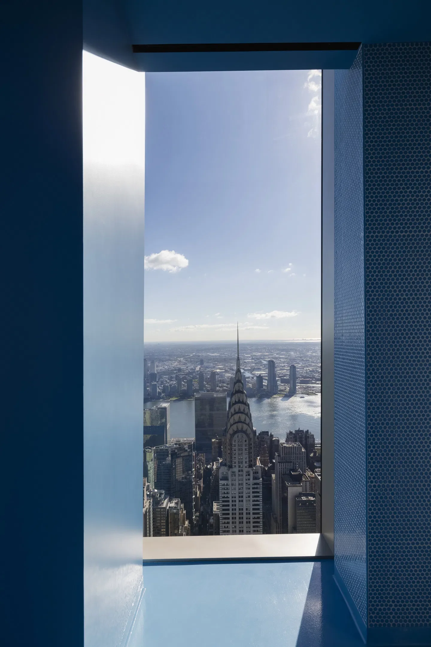 View of the Chrysler Building and New York City skyline through a window on a high-floor observation deck, with blue walls and a clear sky.
