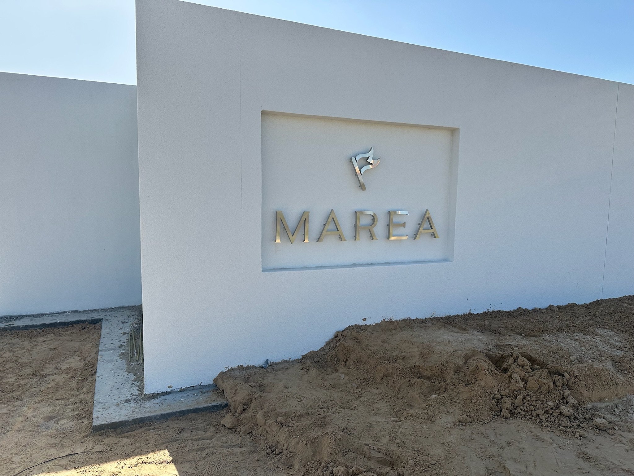 A white wall with a recessed panel displaying a metallic sign with the word "MAREA" and a stylized wave logo above it, under a clear blue sky. There is dirt in front of the wall.