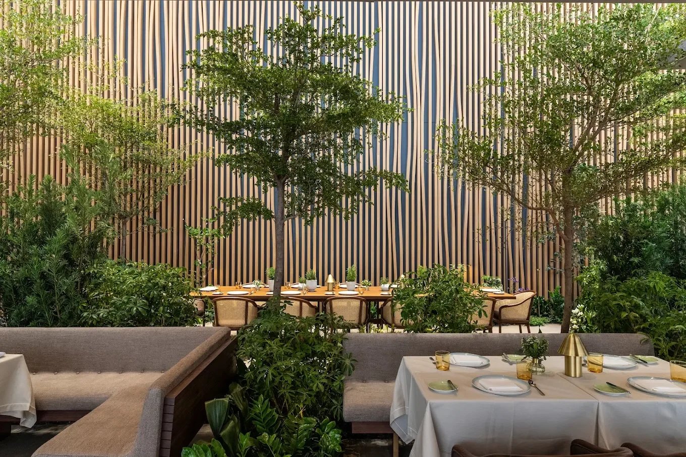 Interior of a restaurant with lush greenery and trees along the wall, dining tables with white tablecloths, gold accents, and place settings.