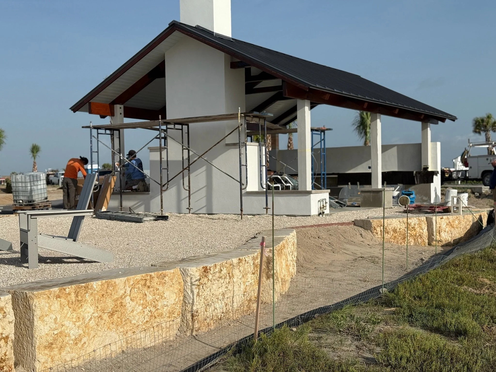 Construction site of a modern house with scaffoldings, workers, and building materials, with palm trees in the background.