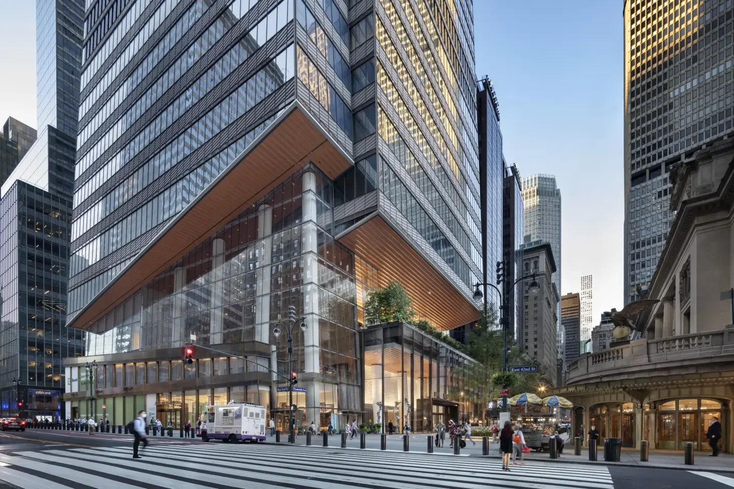 A busy city street intersection surrounded by tall glass skyscrapers with pedestrians crossing and vehicles moving.