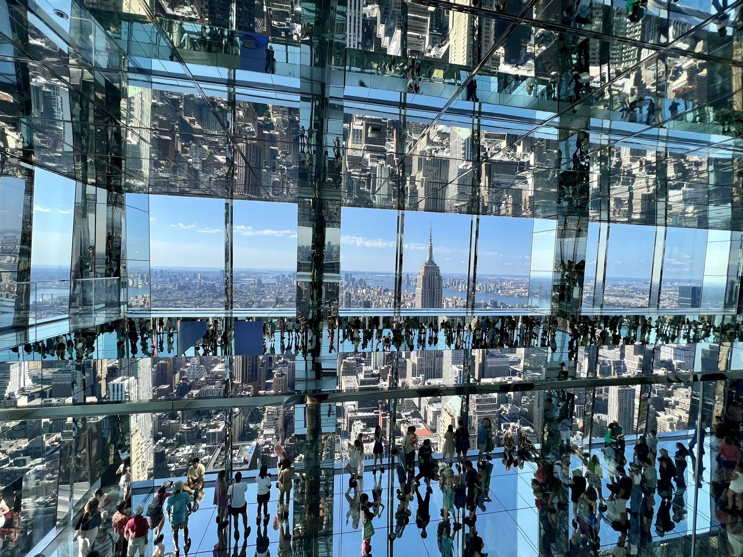 View from 102nd floor observatory of a skyscraper, showing the Empire State Building in New York City through mirrored glass walls with multiple visitors inside.
