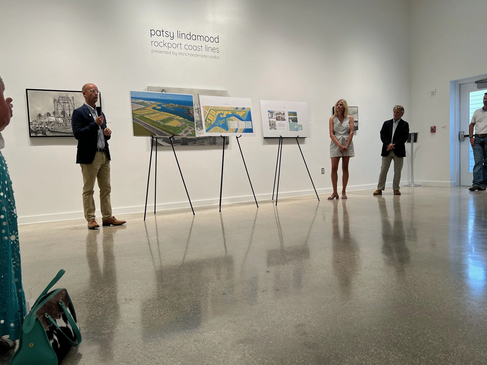 People standing and listening to a speaker at an art exhibit. The exhibit features architectural designs and artwork related to Patsy Lindamood and Rockport Coast Lines.