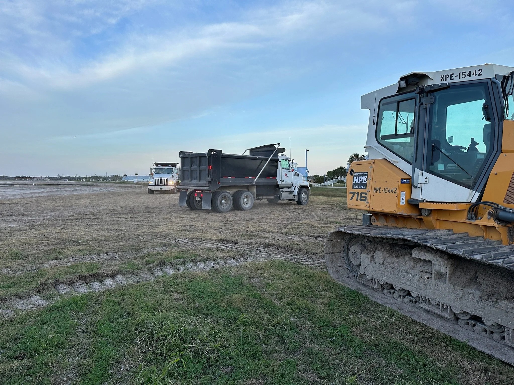 Construction site with heavy machinery, including a yellow excavator in the foreground, a dump truck and a cement mixer in the background on a grassy area near water with a clear sky.