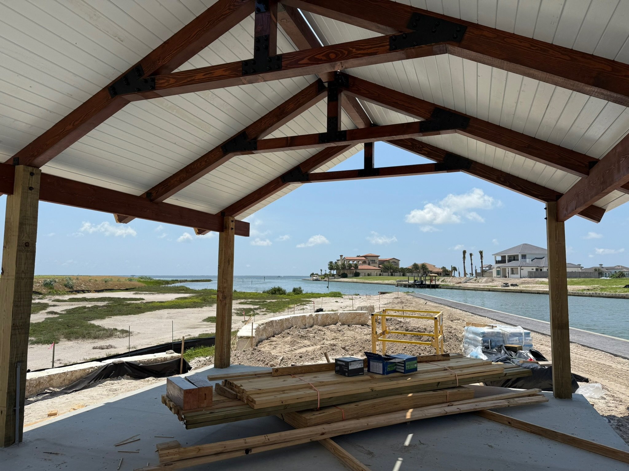 Construction site near a body of water with a partially built wooden structure with a white ceiling. Piles of wood, construction materials, and tools are in the foreground, with a view of houses, waterway, and blue sky with clouds in the background.