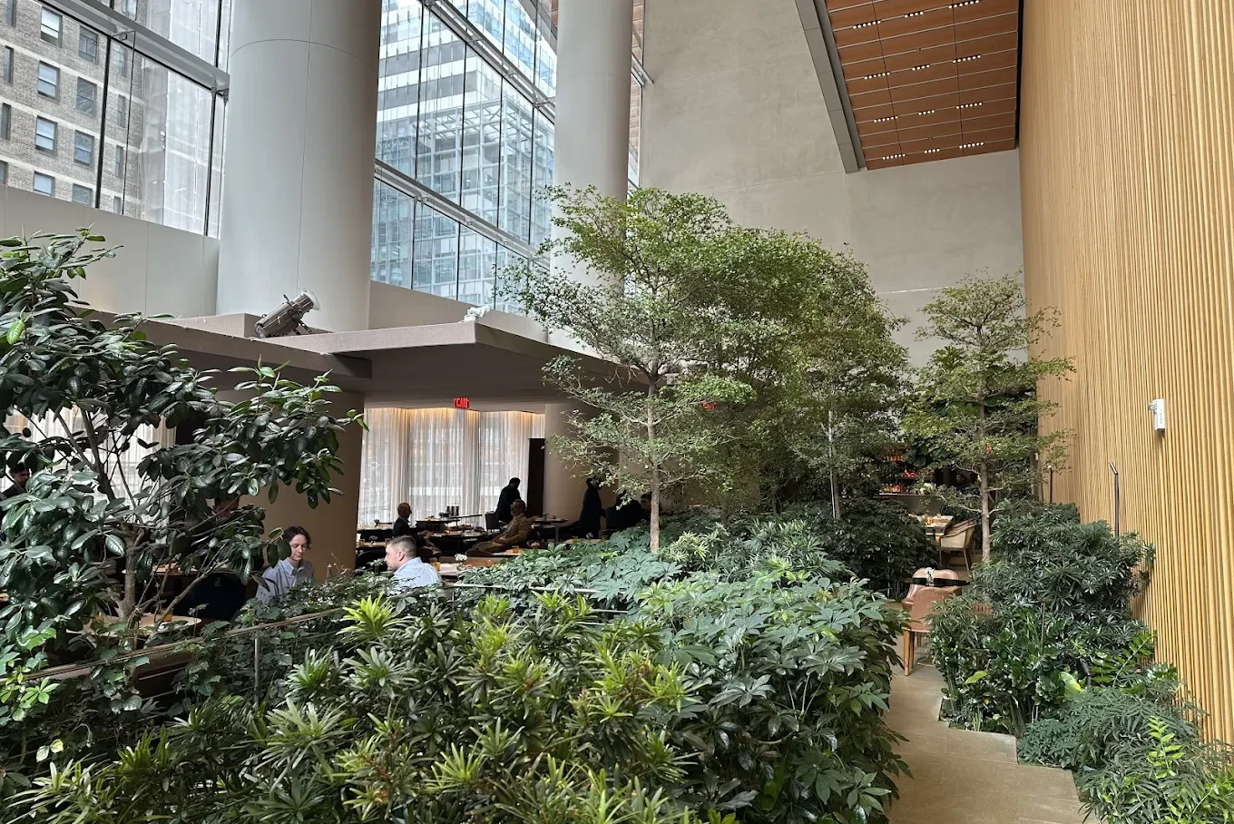 Interior of a modern building with large glass windows, green plants, and people sitting at tables, possibly a restaurant or café.