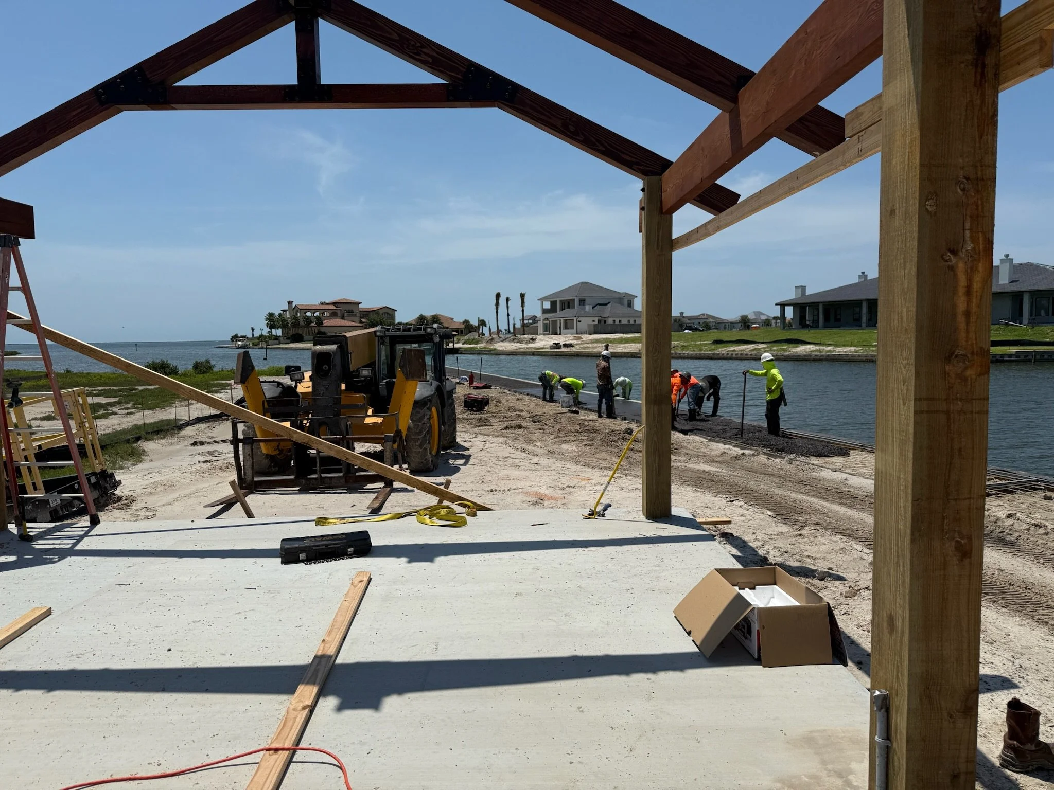 Construction workers building a structure by a waterfront, with houses in the background.