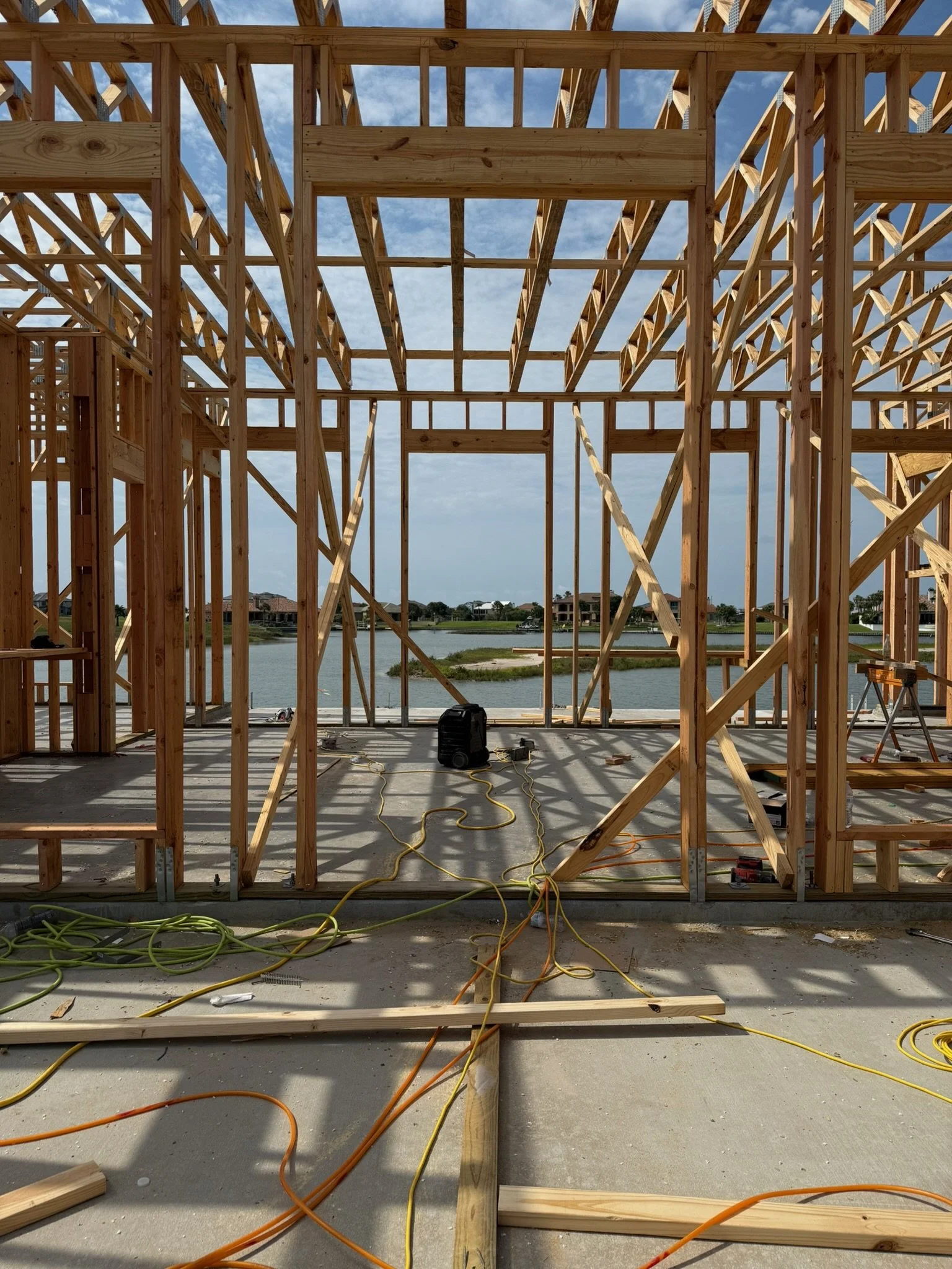 The image shows a wooden framing structure under construction, with various beams and supports. The view extends to a body of water and houses in the background, with a partly cloudy sky above. Construction cords and equipment are present on the grou