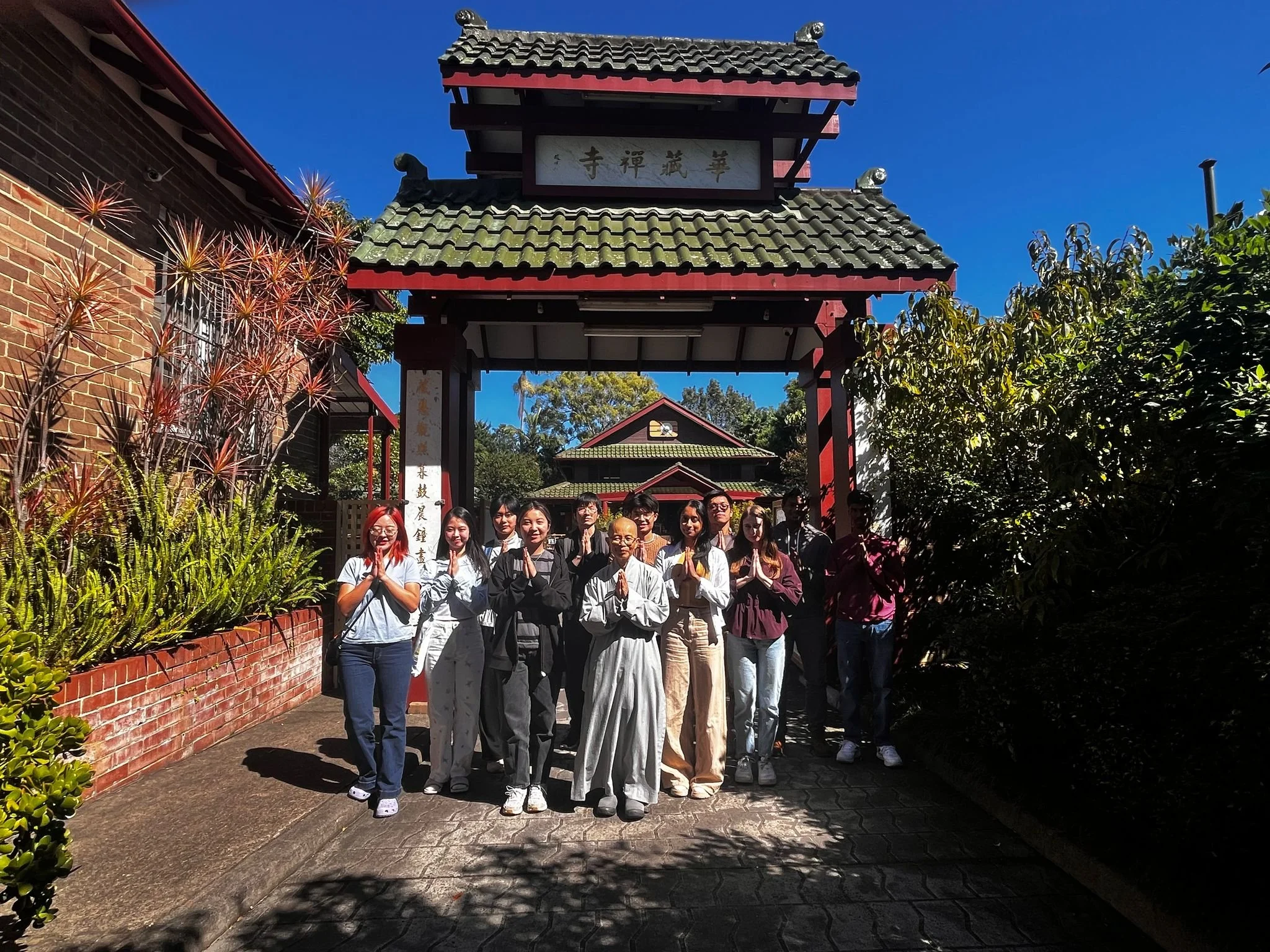 Group of people standing under a traditional Asian entrance gate, with hands in prayer position, in front of a temple or cultural site with a blue sky background.