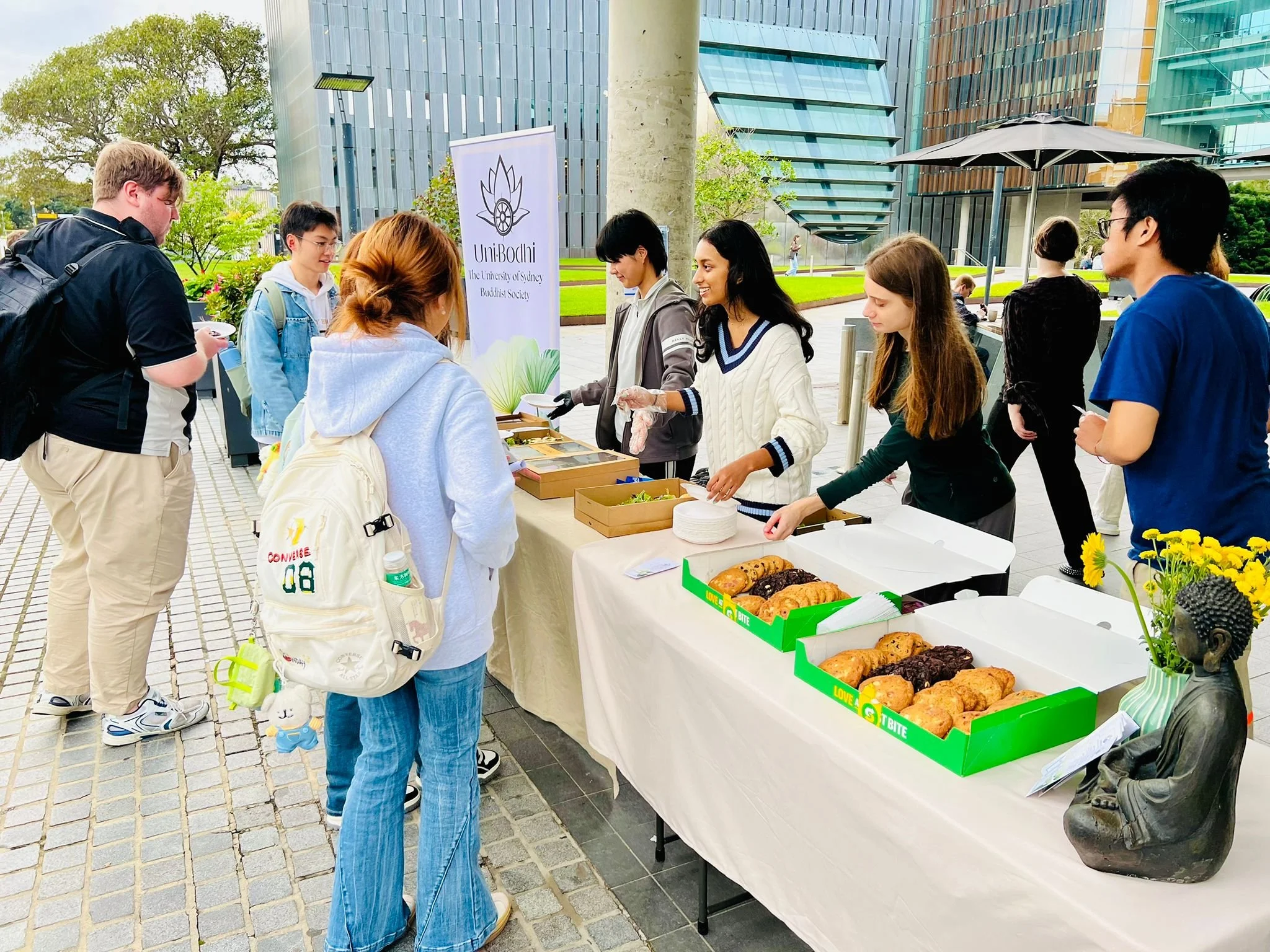 People gather around a table with baked goods at an outdoor event, with a sign for the University of Sydney Buddhist Society in the background.
