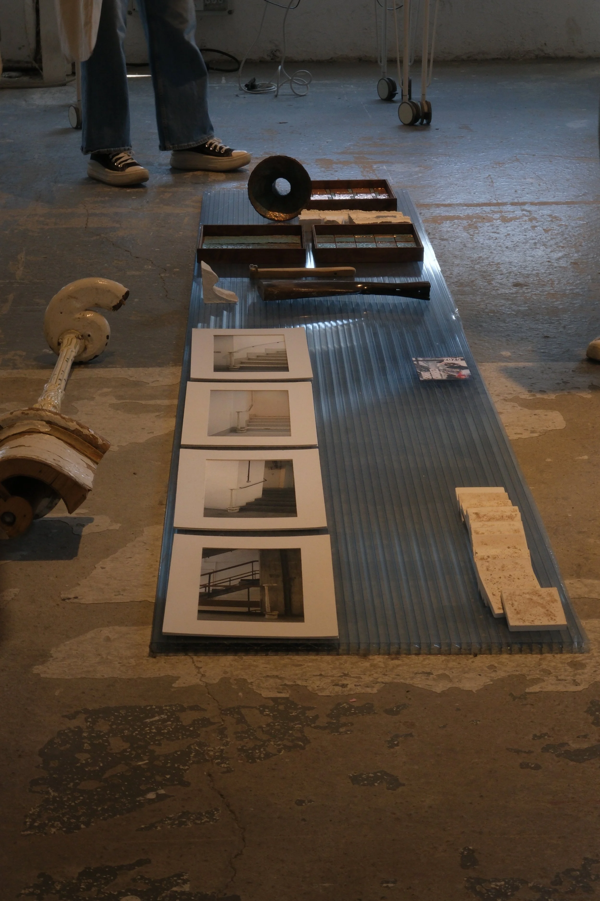 Display of architectural photographs, samples of tile, and spools on a transparent plastic surface on a rough concrete floor.